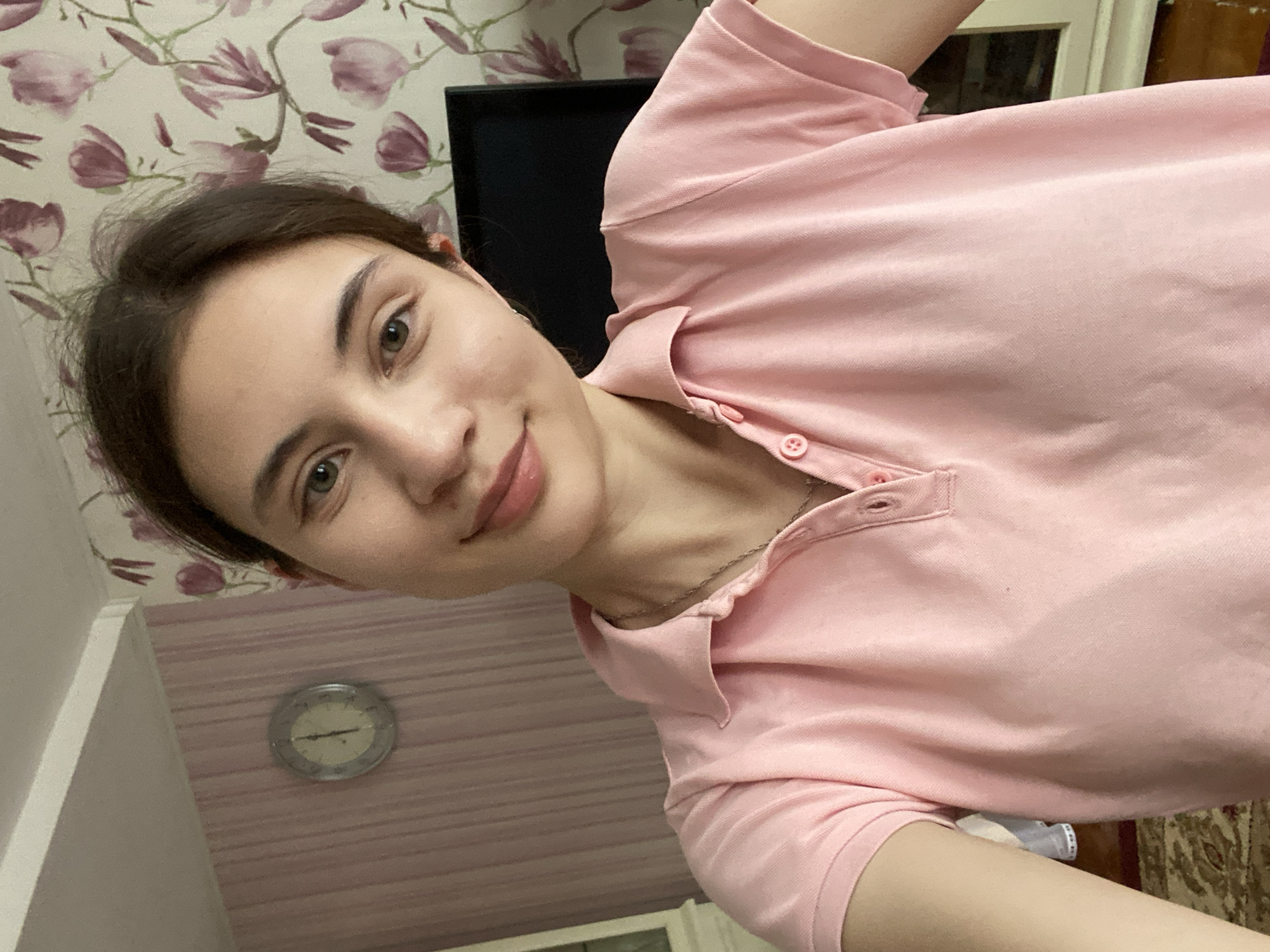 A young woman is taking a selfie in a room. She is wearing a pink polo shirt and a silver necklace. Her hair is brown and pulled back. She has brown eyes and light skin. The room has wallpaper with a floral pattern and a clock on the wall. A television set is visible in the background. The woman is smiling and looking at the camera. The overall lighting is soft and natural. The image is taken indoors, likely in a residential setting. There are no indications of Votkinsk, Russia.