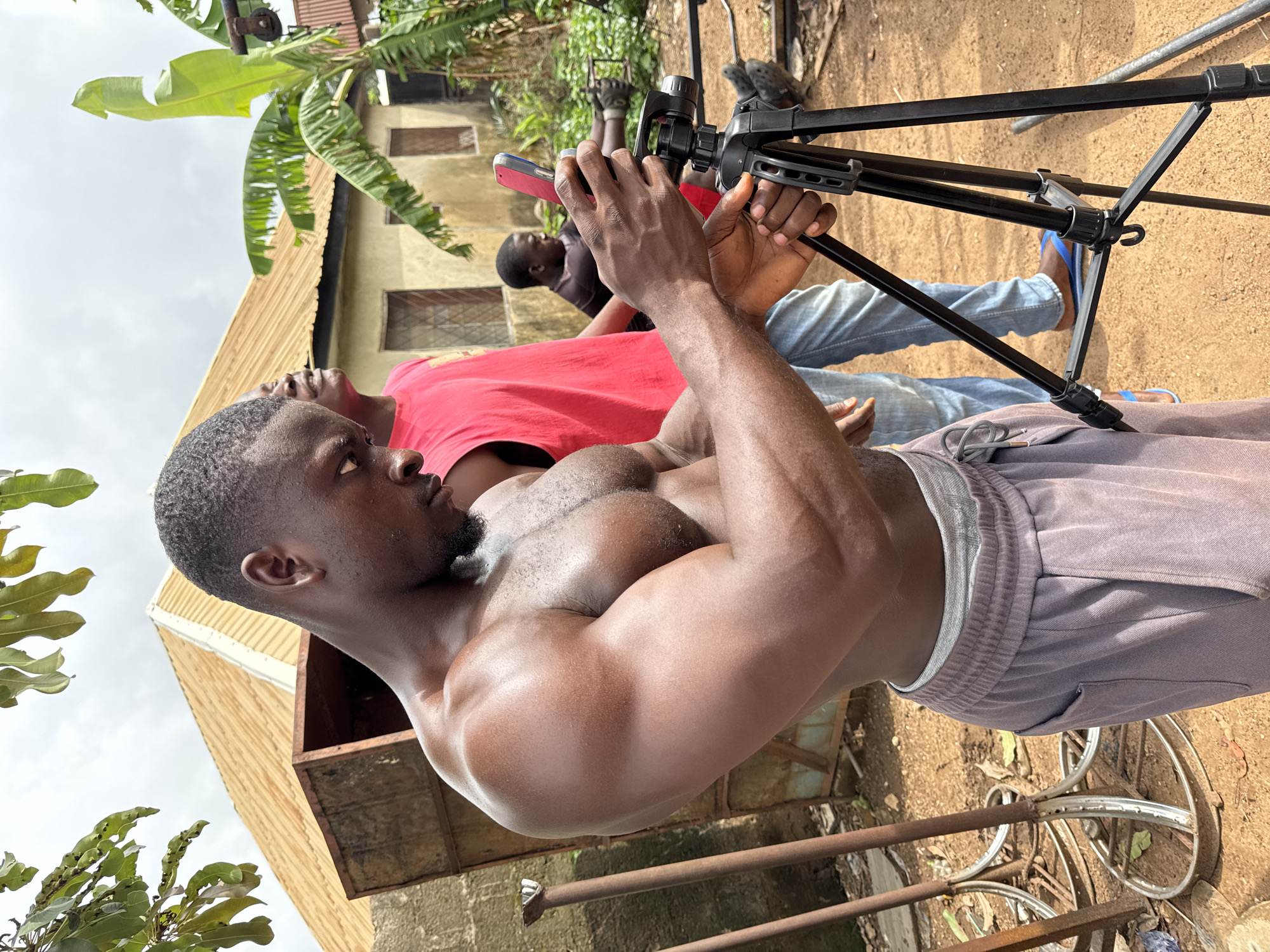 The image shows two men in Akwanga, Nigeria. The foreground figure is a muscular, dark-skinned man with short hair and a beard, shirtless and wearing grey sweatpants. He is focused on adjusting a black tripod in front of him. The man in the background is wearing a red shirt and blue jeans, and he appears to be operating a red device. The ground is dirt, and there's a building with a corrugated metal roof in the background, along with some vegetation. A metal object resembling a bicycle wheel frame lies on the ground near the foreground man.