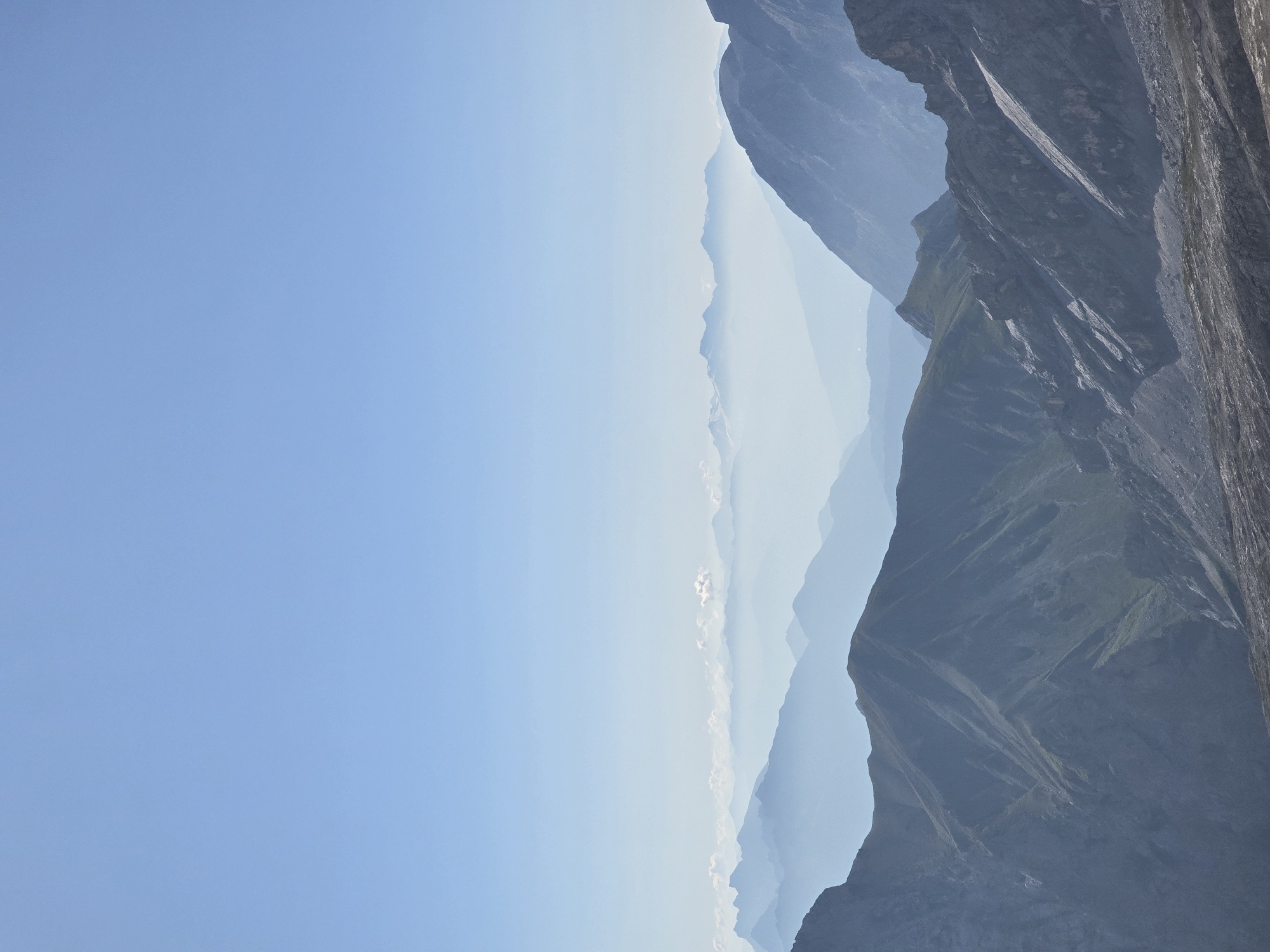 This is a landscape photograph of mountains under a clear blue sky. Multiple layers of mountain ranges recede into the distance, creating a sense of depth and atmospheric perspective. The mountains in the foreground are rocky and detailed, with shadows and highlights defining their rugged terrain. Further back, the mountains become softer and bluer due to atmospheric haze. The sky is a uniform, pale blue, with no clouds visible except for a few wispy formations in the far distance. The scene evokes a sense of vastness and tranquility. While Garmisch-Partenkirchen is known for its mountainous terrain, this particular image does not contain any specific identifiable landmarks that would allow for precise location identification within the city.