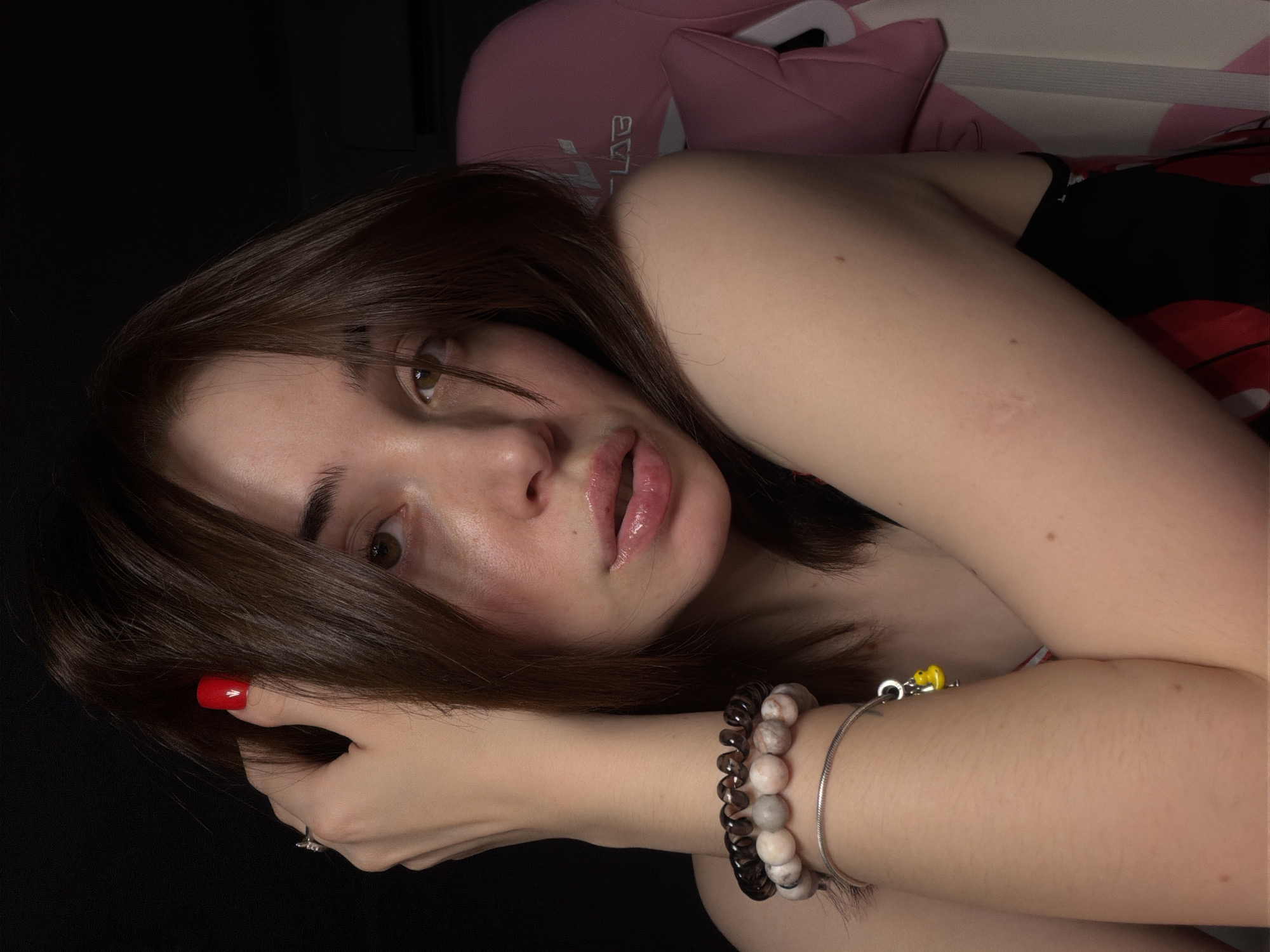 A close-up, medium shot captures a young woman from the chest up, her head tilted slightly as she looks towards the camera. She has medium-length brown hair that falls loosely around her face, with a strand obscuring part of her left eye. Her right hand is raised, fingers brushing through her hair, and her fingernails are painted a vibrant red. She wears a silver bracelet on her left wrist, adorned with dark, banded beads and a small yellow charm. Her lips are parted, and they appear glossy and slightly flushed, as if she is wearing lip gloss or balm. Her skin is fair, with subtle pink blush on her cheeks. In the background, a pink gaming chair is partially visible. The lighting is dim, creating a soft, intimate atmosphere. The background is dark and out of focus.