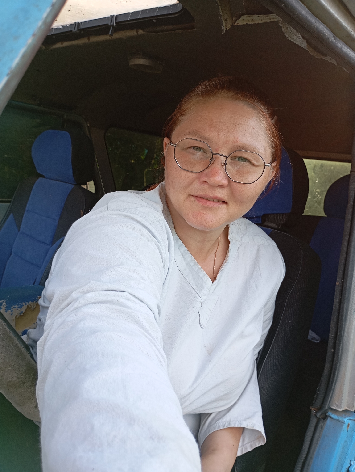 A woman with reddish hair, wearing glasses and a white shirt, is taking a selfie inside what appears to be a vehicle. She is seated in the driver's seat, with the passenger seat visible behind her. The interior of the vehicle shows blue and black upholstery on the seats and a worn blue painted dashboard. The exterior of the vehicle is a dull blue. The background outside the vehicle is blurred but suggests a natural, possibly rural, setting with greenery. The lighting indicates it is daytime, likely sunny. The woman's expression is neutral, and she appears to be looking directly at the camera. The overall impression is a candid, informal self-portrait taken in a vehicle in what could be a rural area of Ryabki, Russia. There is no visible text in the image.