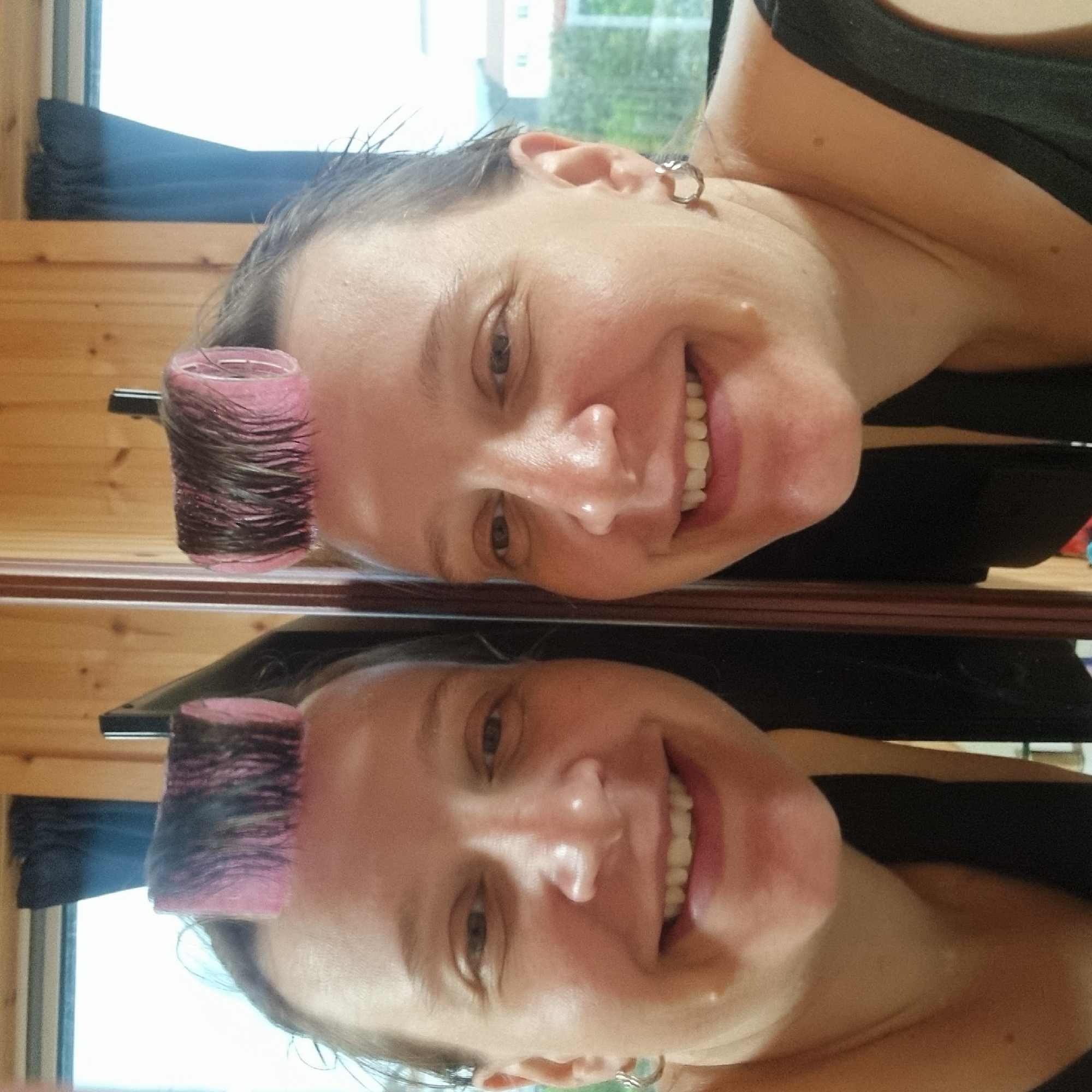 The image is a selfie of a woman with pink hair rollers in her hair, taken indoors in a room with wood-paneled walls. She is smiling and looking at the camera. The room appears to be a bedroom or bathroom, with a window visible in the background. The woman is wearing a black top. The image is a reflection in a mirror, showing her from the chest up.
