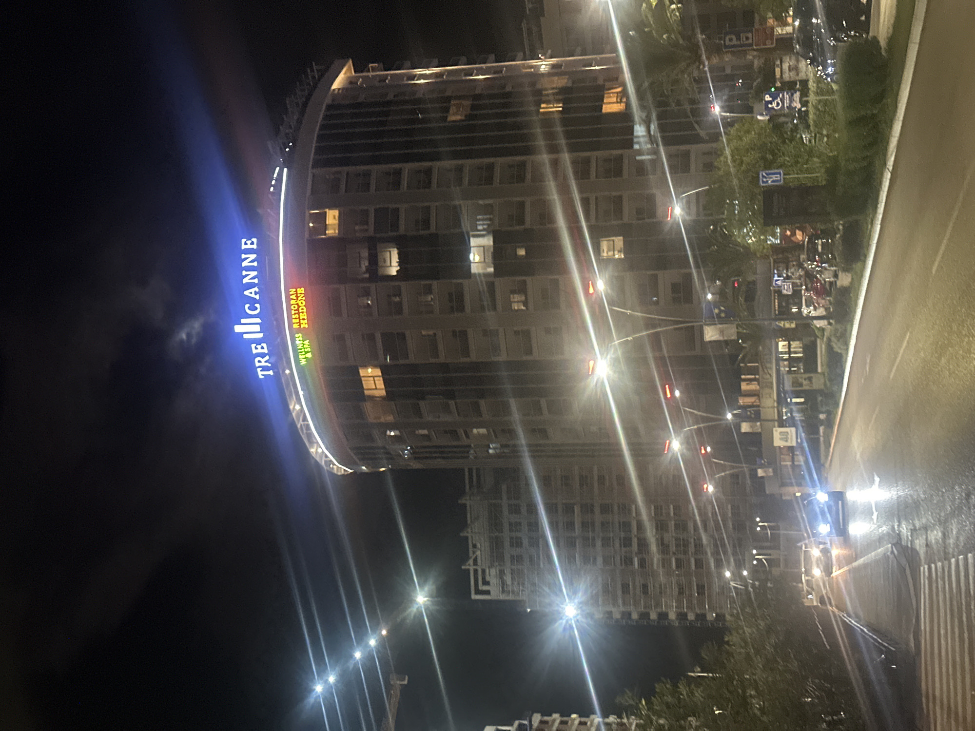 The image shows the Tre Canne hotel complex in Budva, Montenegro, at night. The hotel's curved facade is illuminated with the name "TRE CANNE" in blue neon lights. Adjacent signs are visible, indicating "Wellness Restoran & Spa Hedone." The building is a multi-story structure with numerous windows, some illuminated. Streetlights line the adjacent road, which reflects the light, indicating wet conditions. There are also trees and greenery present along the road. In the distance, another building is visible, also lit. Traffic signs and cars can be seen on the street. The sky is dark, suggesting nighttime.