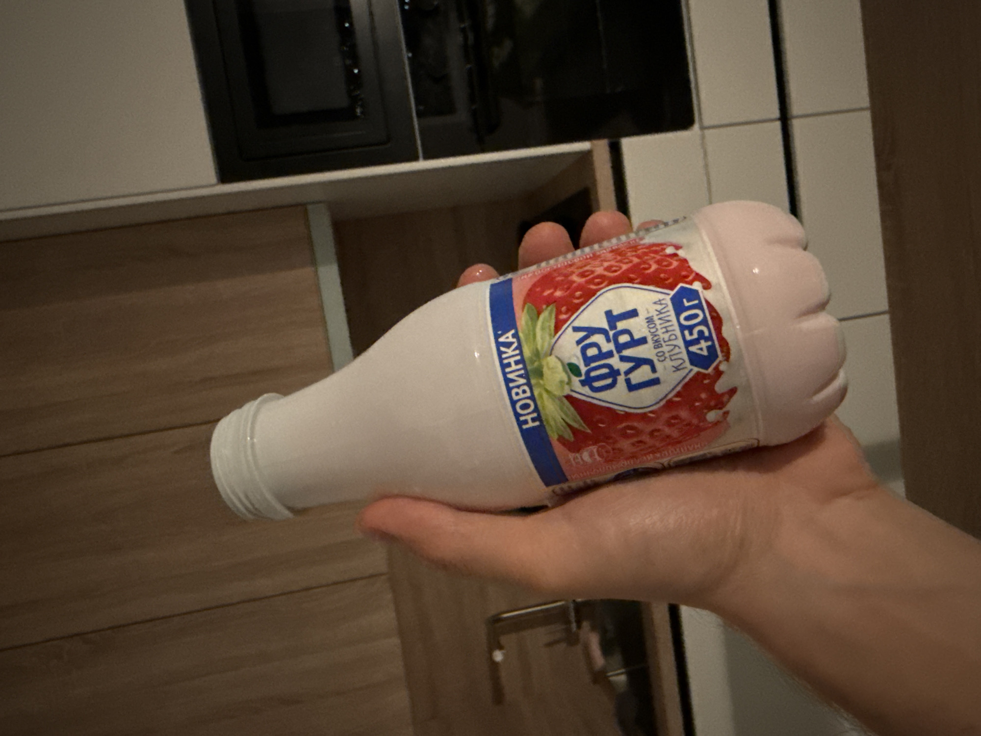 A person is holding a plastic bottle of strawberry-flavored yogurt in a kitchen. The bottle has a white cap and a pink liquid inside. The label on the bottle is in Russian and features an image of a strawberry. The kitchen appears to be modern, with wooden cabinets and a black microwave in the background. The lighting is dim, suggesting it might be evening. The background also shows white tiled surfaces and wooden flooring.