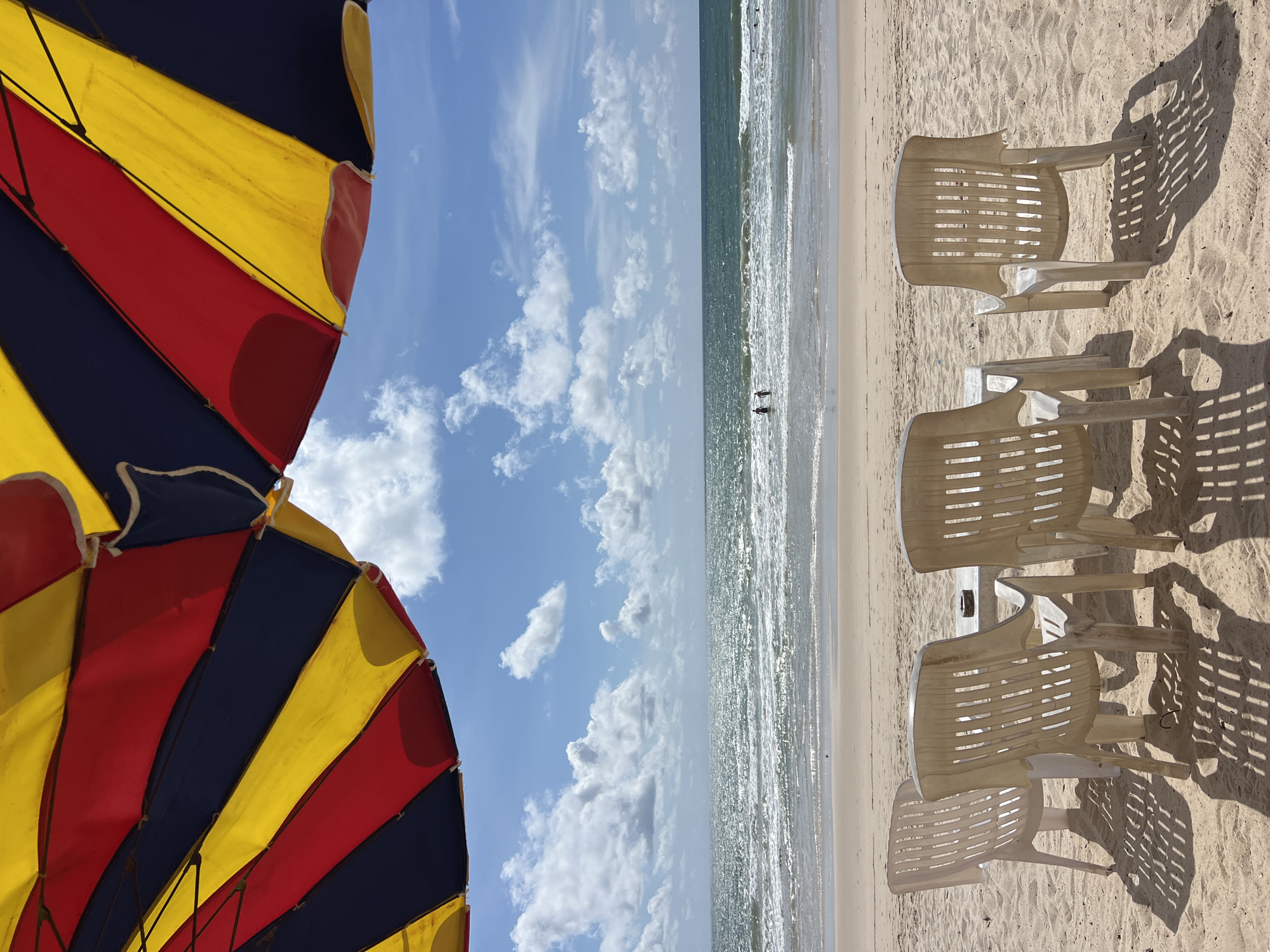 A beach scene in Choeng Thale, Thailand, features a colorful umbrella providing shade over a table and four white plastic chairs arranged on sand. In the background, the ocean stretches to the horizon under a bright blue sky with scattered white clouds. Two individuals are visible in the shallow water, wading near the shore.