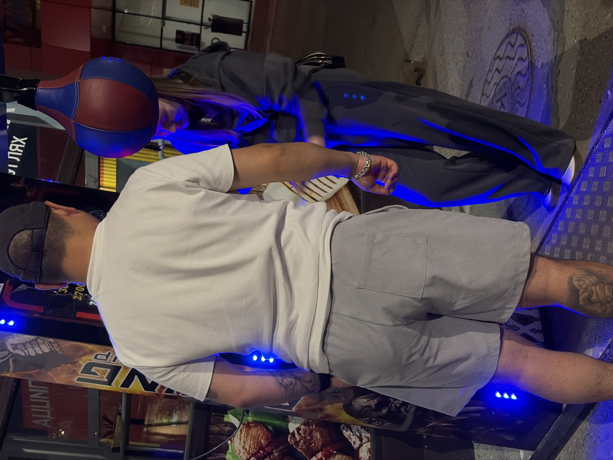 Two individuals, a male and a female, are standing next to a boxing apparatus in what appears to be an outdoor entertainment area. The male, seen from the back, is wearing a white t-shirt, grey shorts, and a dark baseball cap. He has tattoos on his arm and calf. The female, facing away from the camera and looking down, is wearing a dark hooded sweatshirt and dark pants. She is standing near a punching bag that is suspended from a metal frame. Blue LED lights are illuminating the scene. The background suggests an urban setting with various signs and structures, possibly a street or a park area. The overall atmosphere is that of a casual nighttime setting. The location is identified as Chaikovsky, Russia.