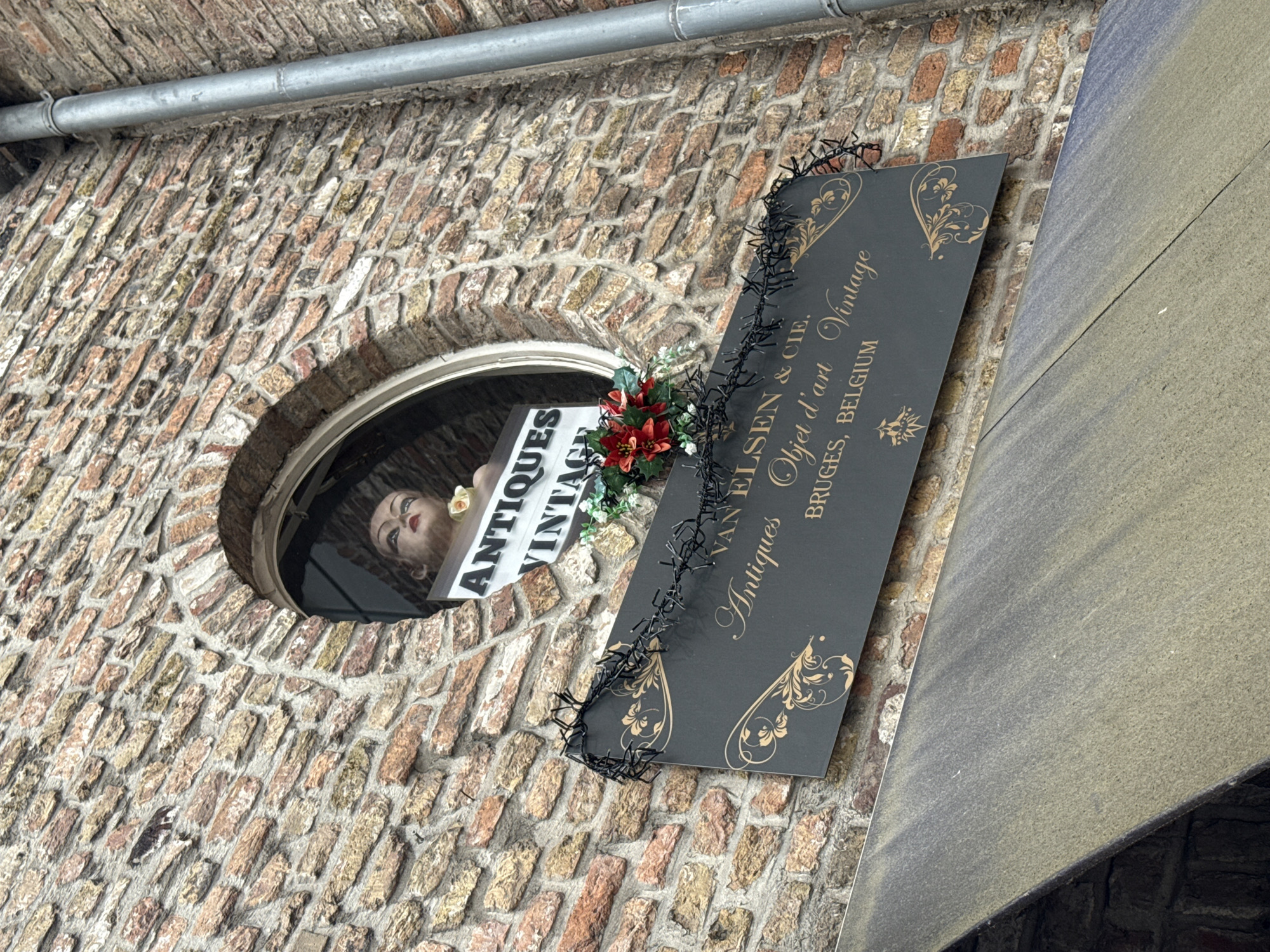 An outdoor, eye-level, medium shot captures a brick building facade in Bruges, Belgium. A round window on the second story displays a mannequin head with makeup, and a sign that reads "ANTIQUES VINTAGE". Below the window, a dark rectangular sign is adorned with black string lights and a floral arrangement. The sign states "VAN ELSEN & CIE.", "Antiques Objet d'art Vintage", and "BRUGES, BELGIUM". A silver downspout runs vertically along the right side of the building. The lower portion of the image shows a grey, textured awning.