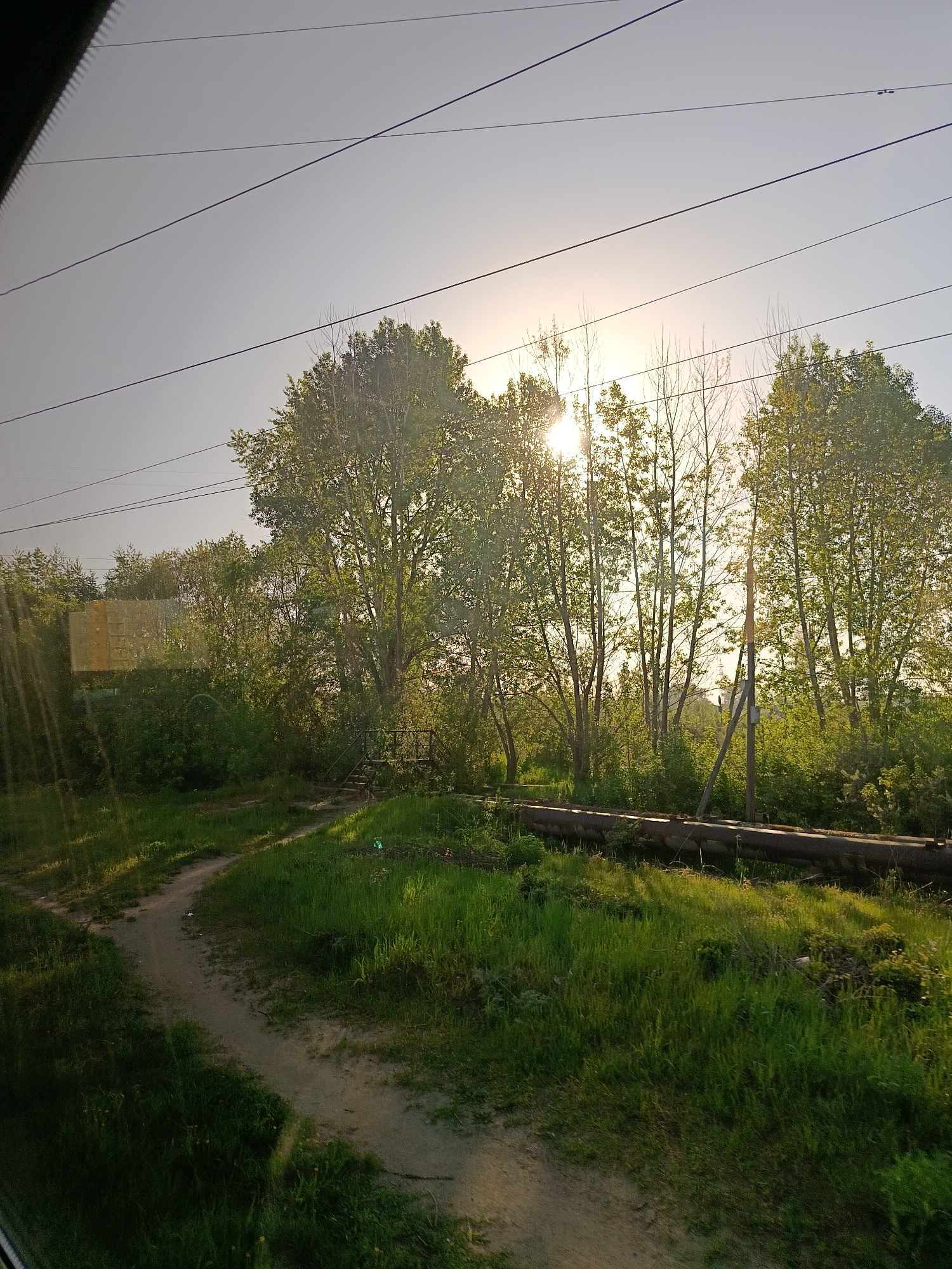 The image shows a view from what appears to be a vehicle window in a natural setting, likely near Chaikovsky, Russia. The sky is bright, with the sun visible through the trees, creating a strong backlight effect. A dirt path curves through green grass in the foreground. A raised pipeline structure crosses the scene. Trees are the dominant feature of the middle ground. Several utility lines run across the top of the frame. The overall environment suggests a rural or suburban fringe of a city, rather than a densely urban setting.