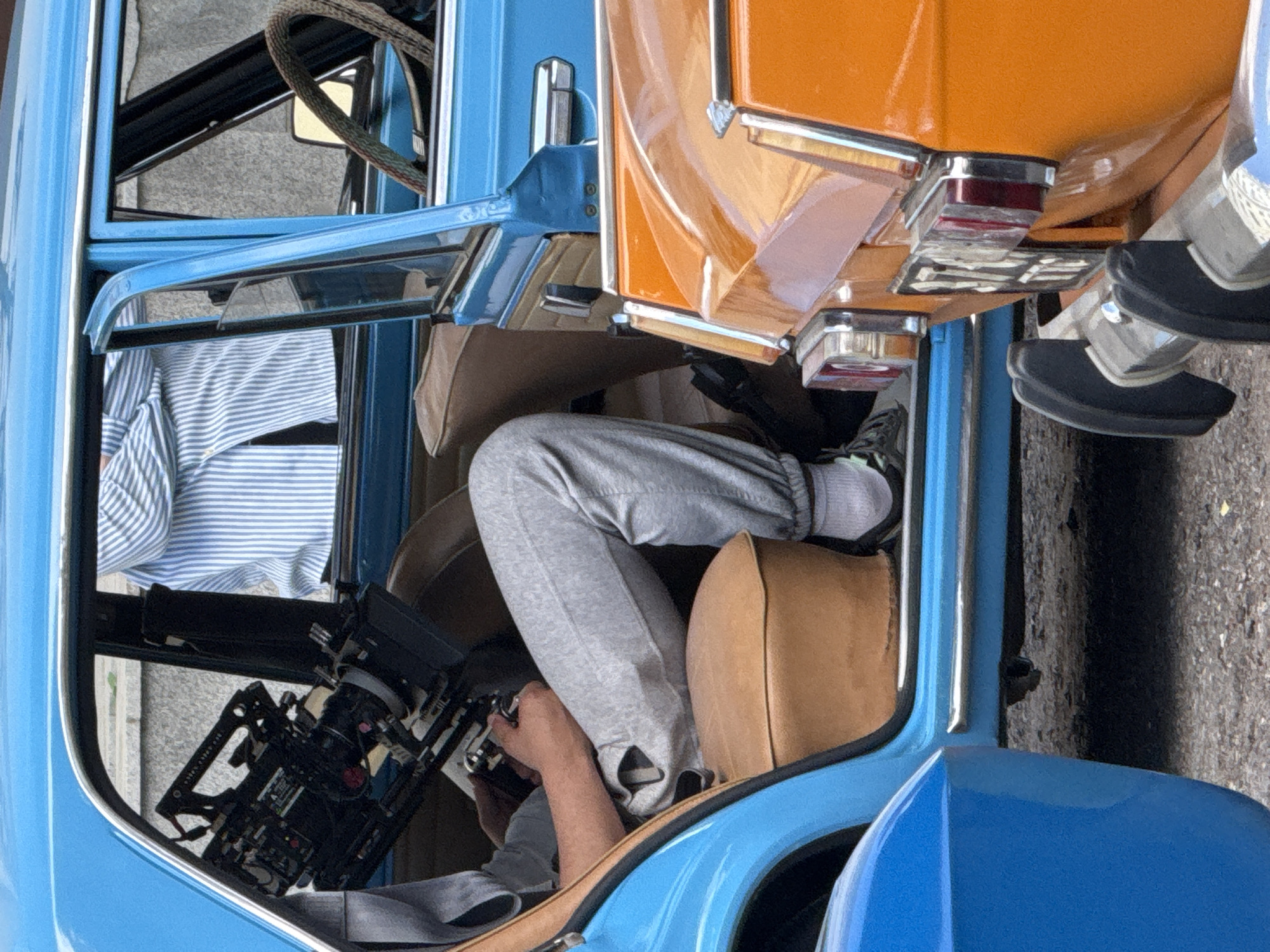 A ground-level, eye-level shot depicts the interior of a vehicle, focusing on the driver. The driver is wearing a blue and white striped shirt, gray sweatpants, white socks, and black shoes, and is sitting on a tan leather seat. The person is operating a camera and is positioned behind the steering wheel. To the right of the vehicle, an adjacent vehicle is visible, painted orange and blue. The location is speculated to be Kazan, Russia.