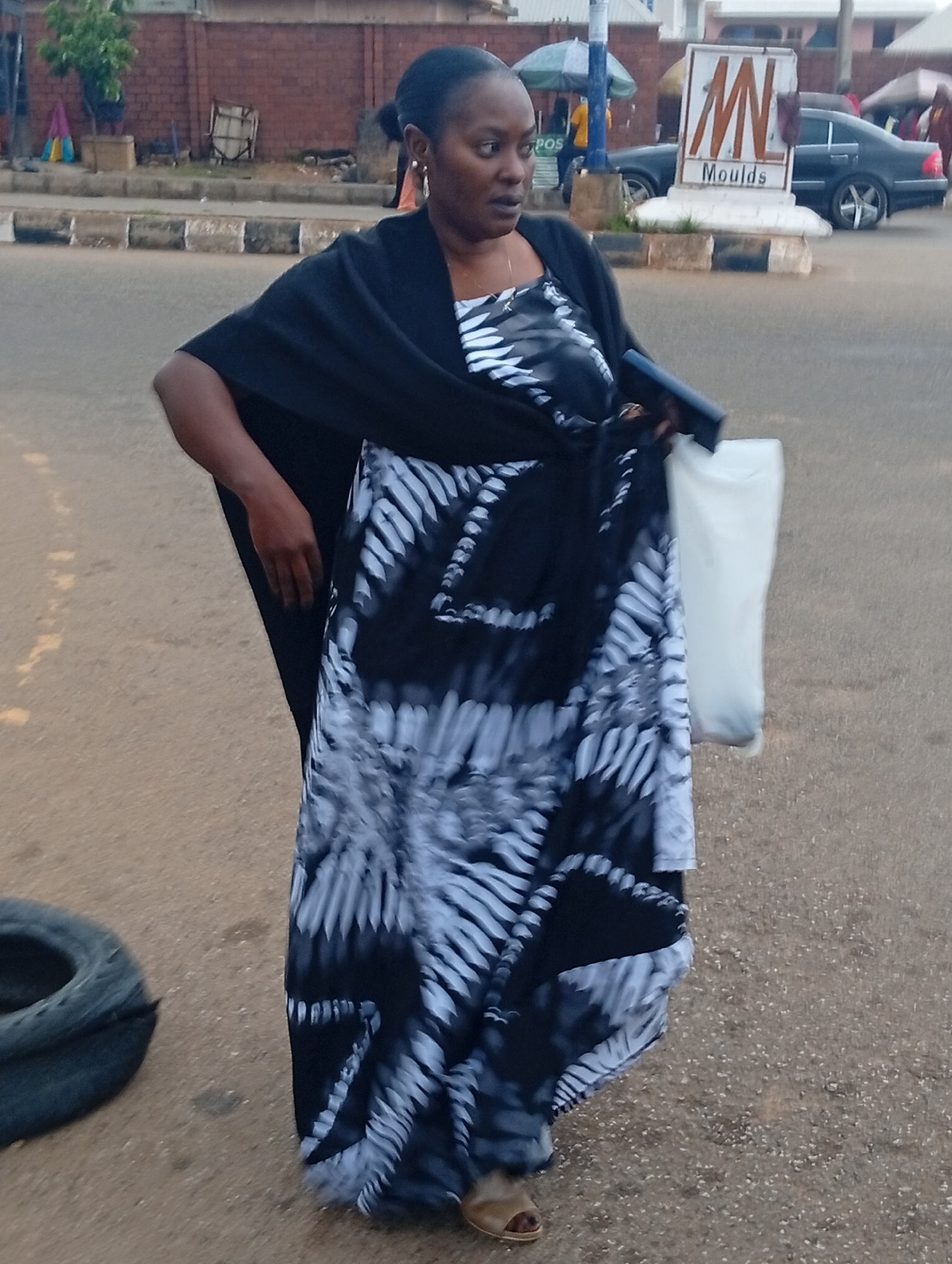 In Jos, Nigeria, a Black woman stands on a paved street, holding a white plastic bag and a dark object in her hands. She is wearing a black shawl draped over a black and white patterned dress. She is also wearing earrings and sandals. A car, a sign that says "MN Moulds", and other people are visible in the background. The scene appears to be a street in a commercial area.