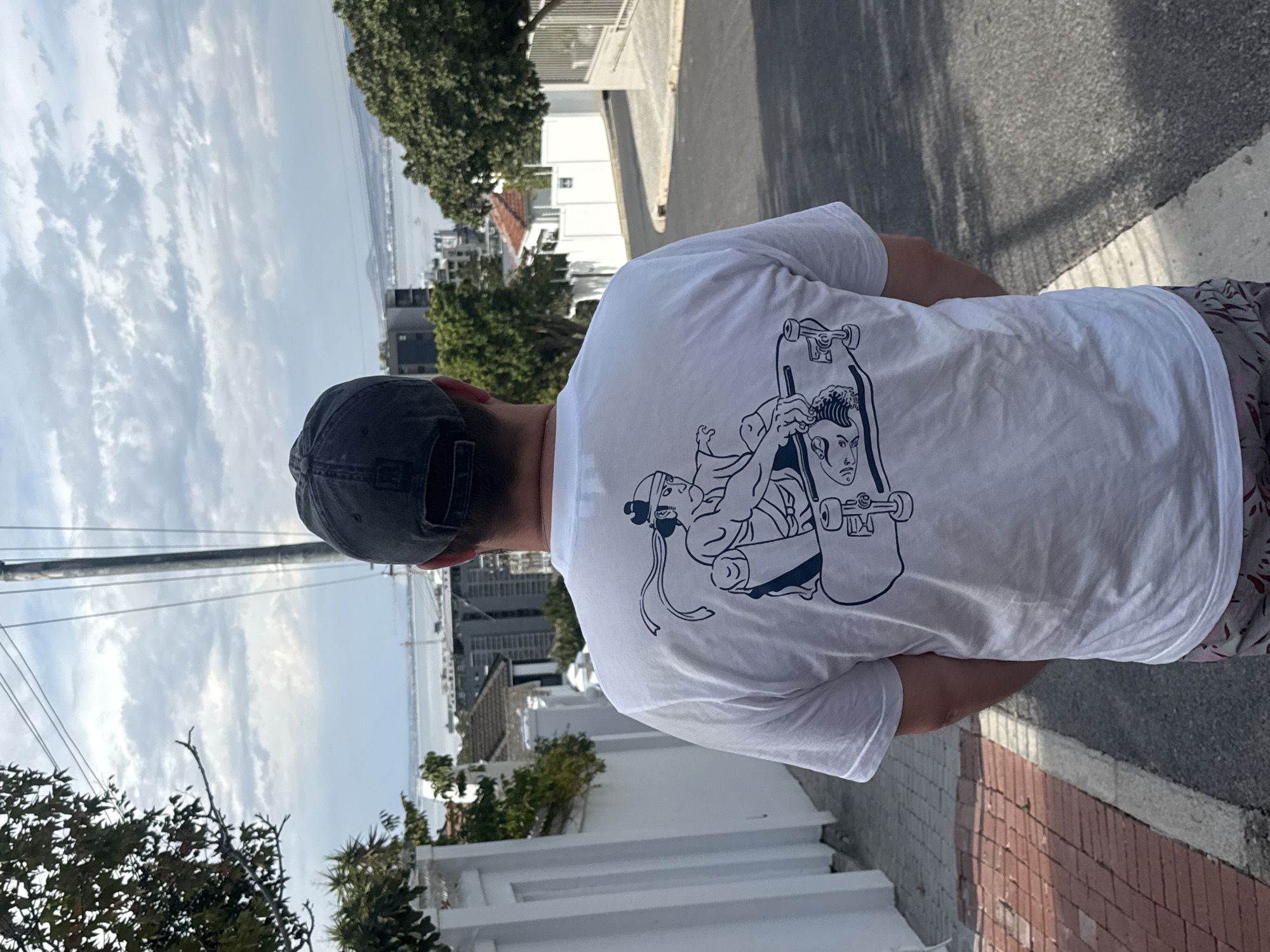 Here is a detailed description of the image:
The image is taken outdoors and depicts a person from the back, wearing a white t-shirt with a graphic print, a dark baseball cap, and printed shorts. The person is standing on a sidewalk made of red and grey bricks.
In the background, there is a cityscape with buildings, trees, and a body of water. The sky is overcast with clouds. The location is likely Cape Town, South Africa, due to the urban setting and the visible coastline.