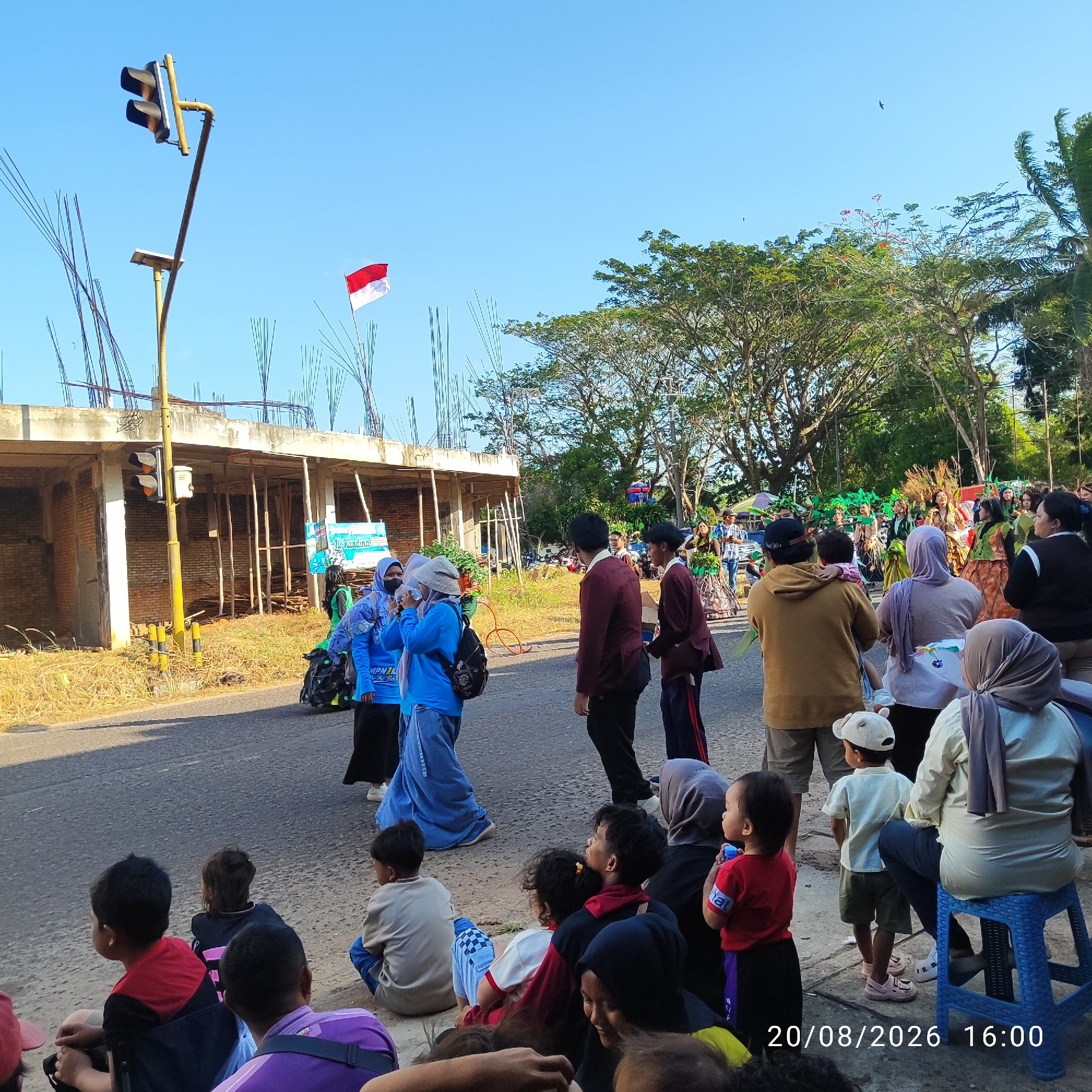 In Buding, Indonesia, on August 20, 2026, at 16:00, a street scene unfolds under a clear blue sky. A crowd has gathered, seated and standing along the side of a road, facing a street lined with trees and construction. In the background, a building under construction, with exposed brick and scaffolding, is visible. A traffic light stands tall on a yellow pole. A red and white Indonesian flag flutters in the breeze. People, including adults and children, are dressed in a variety of clothing, some in traditional attire with vibrant colors and headscarves, others in casual wear. Some individuals appear to be observing an event, possibly a parade, as indicated by the colorful costumes and the gathering of people. The atmosphere appears festive and communal, with people engaging with each other and enjoying the occasion.