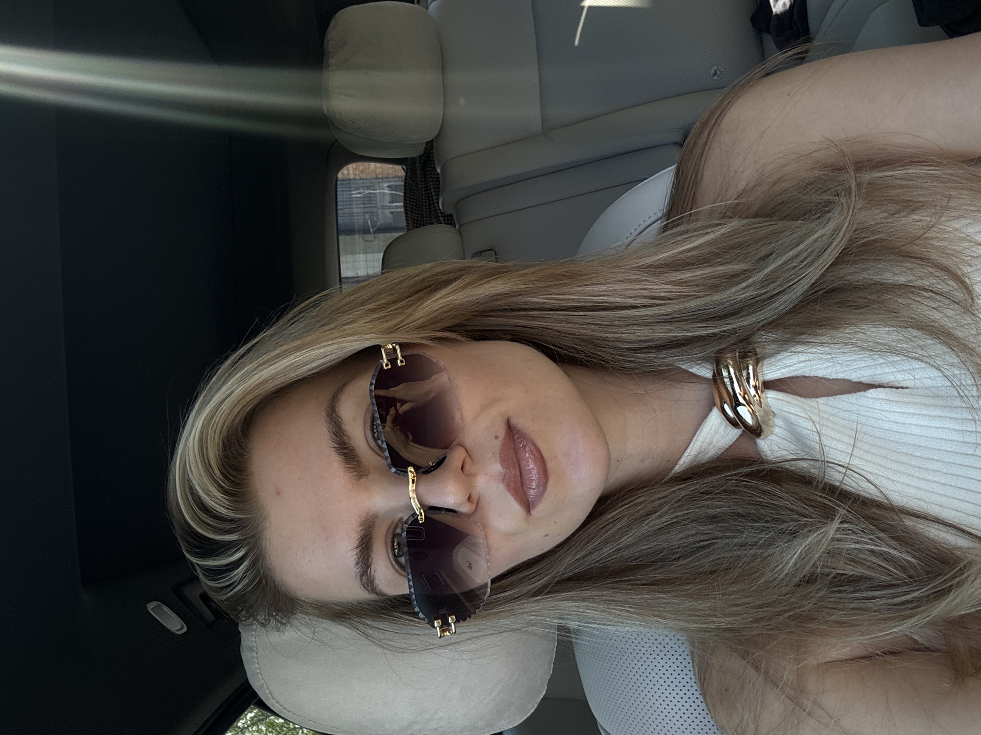 A woman with blonde hair and sunglasses is sitting in a car. She is wearing a white top and a gold necklace. The car has light-colored seats and a black interior. The location is Unknown, Unknown.