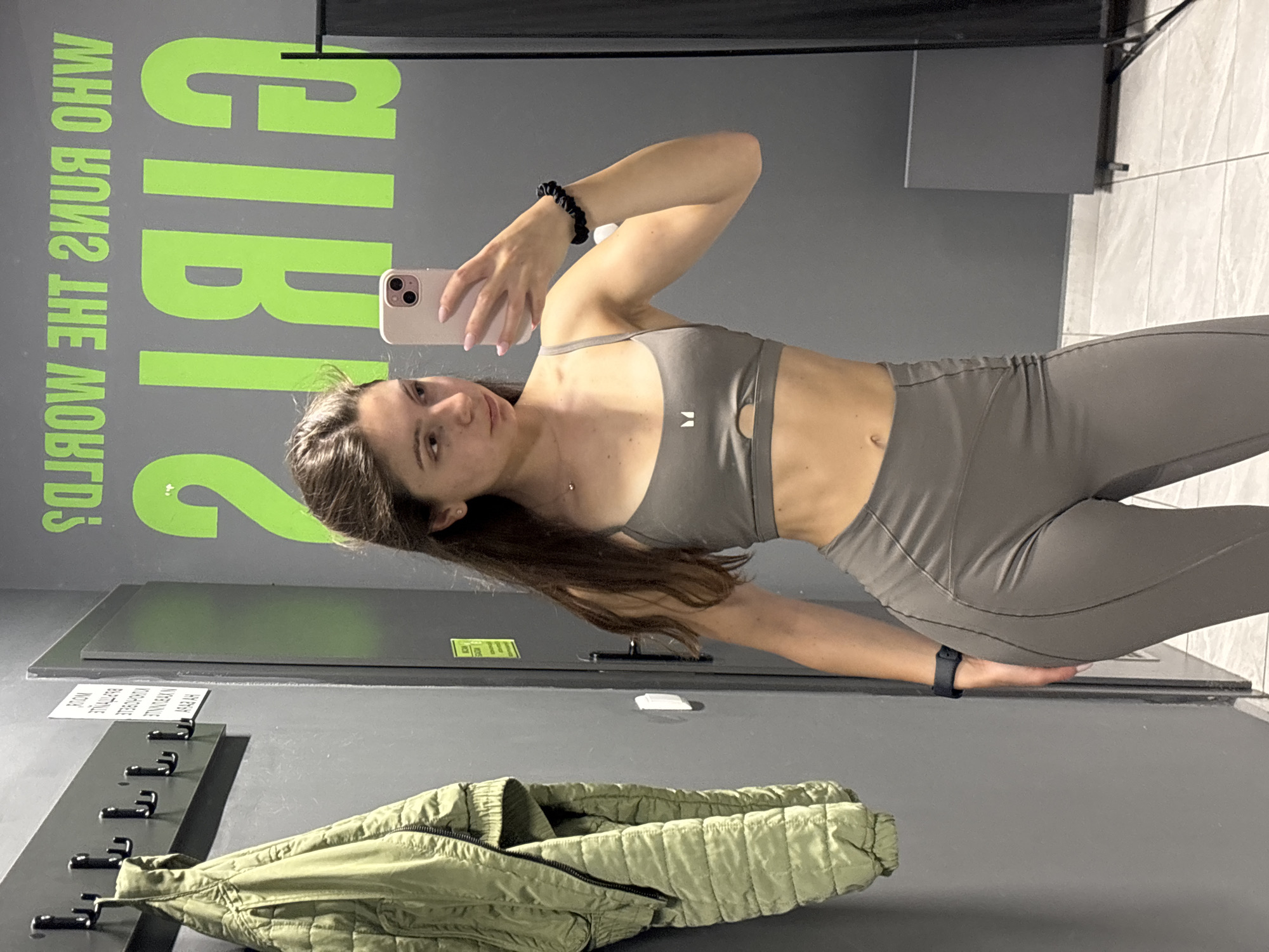 A young woman with long, dark hair is taking a mirror selfie in what appears to be a gym locker room. She is wearing a grey sports bra and matching leggings. A green jacket is hanging on a rack to her left. The wall behind her has large, bright green lettering that reads "GIRLS WHO RUN THE WORLD" in reverse. There are also lockers and a trash can visible in the background. The floor is tiled. There is no information to suggest the location is in Varna, Bulgaria.