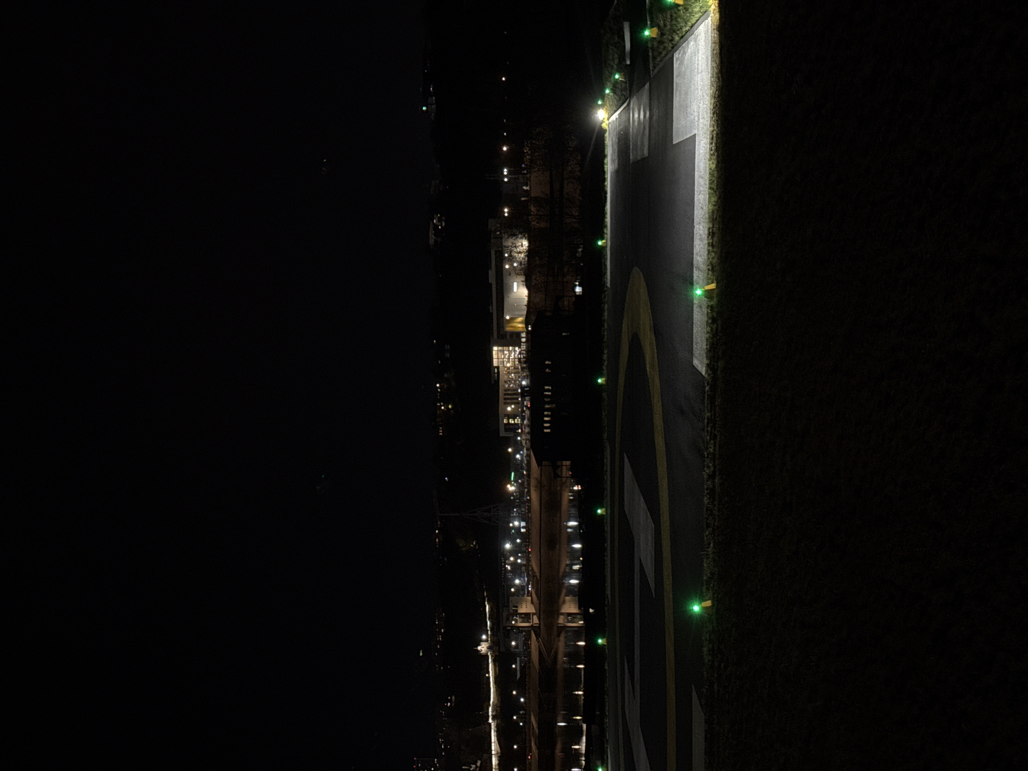 The image shows an aerial view at night in Nizhny Novgorod, Russia. A helipad is in the foreground with green lights marking its perimeter. Beyond the helipad, there is a waterway with city lights reflected on its surface. Further into the distance, buildings and streetlights are visible in the cityscape.