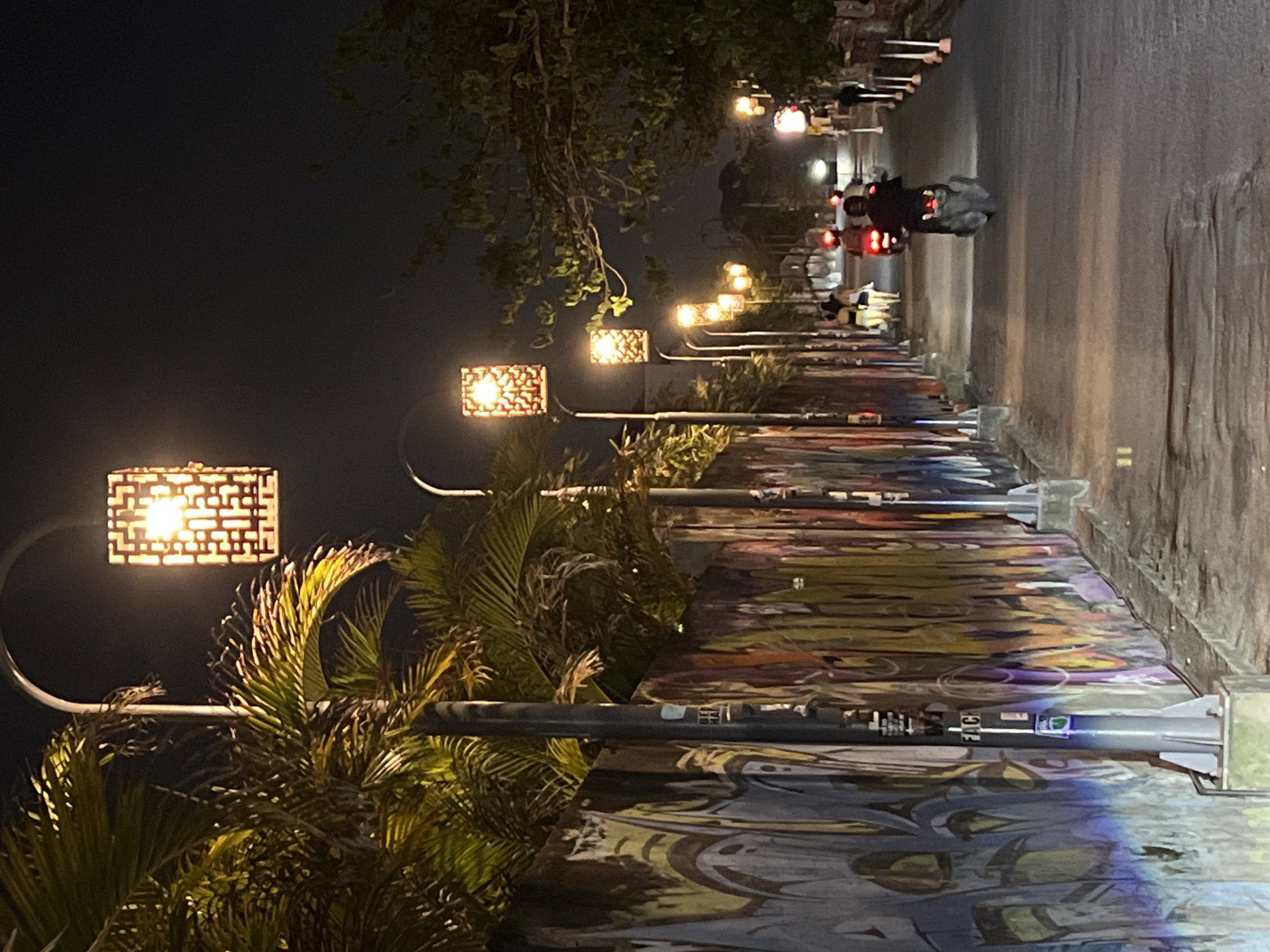The image shows a street scene in Canggu, Indonesia, at night. The street has a sidewalk lined with a mural and illuminated by decorative street lamps. A motorcycle with two riders is moving along the road, away from the camera. Several pedestrians are walking on the sidewalk in the distance. Palm trees and other vegetation are visible along the edge of the street, partially obscuring some of the street lamps.