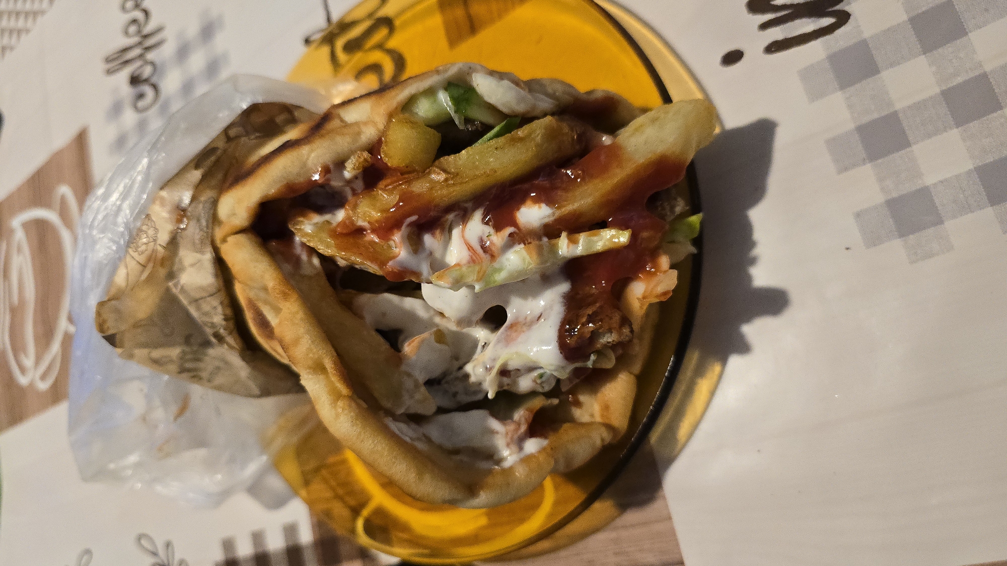 A close-up, overhead shot shows a gyro, wrapped in paper and plastic, sitting on a yellow plate. The gyro is filled with meat, fries, lettuce, and sauce. The plate is on a tablecloth with a coffee cup design and a checkered pattern. The background is blurred.