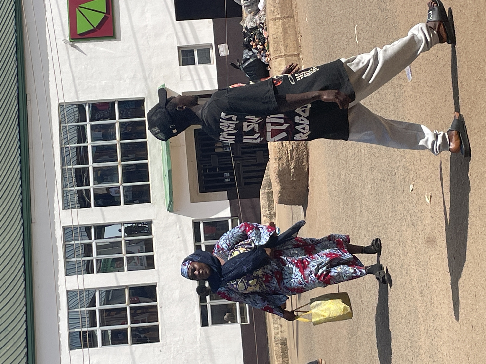 In the city of Bukuru, Nigeria, a woman in a blue, red, and yellow patterned dress and a dark blue headscarf walks to the left, carrying a yellow shopping bag. She wears dark sandals. A young man, facing to the right and wearing a black t-shirt with white text, a black baseball cap, and light gray sweatpants, walks in the foreground towards the right. He wears black slides. Behind them, a white building with a green roof features several large windows. To the right of the building, black trash bags and scattered debris are visible on the ground. The scene takes place on a paved street under a bright sky, casting long shadows on the ground.