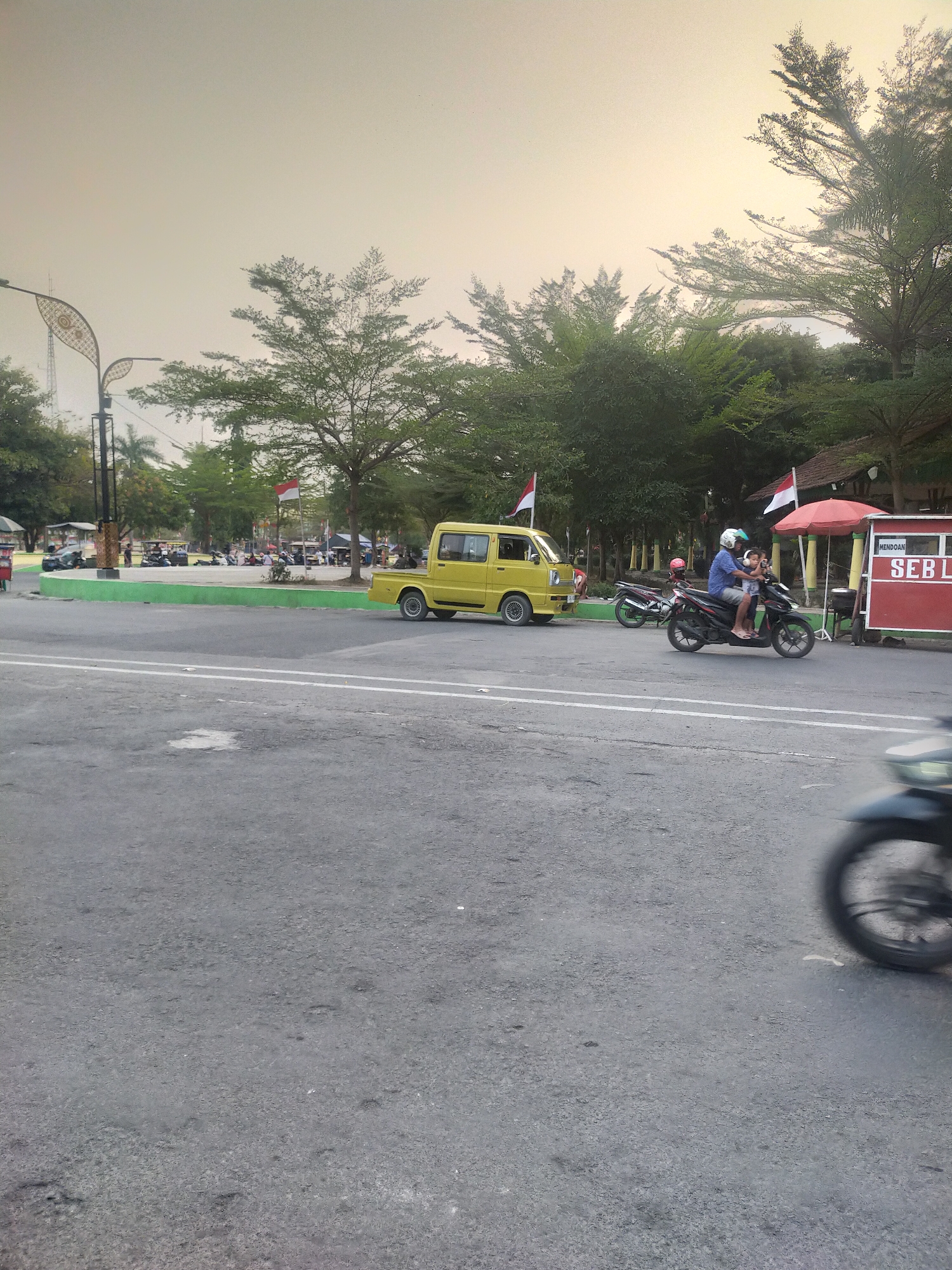 The image captures a street scene in Slawi, Indonesia, under an overcast sky. In the foreground, a paved road with white lane markings dominates the lower half of the frame. To the right, a motorcycle is a blur of motion as it passes by, its wheel visible.
In the mid-ground, several individuals are present, with two on a black motorcycle wearing helmets. One person is driving, and a child is seated in front of them. A yellow pickup truck is parked on the left side of the road. A red and white kiosk is situated on the right, with a red umbrella above it. Indonesian flags, red and white, are visible on flagpoles in the background.
The background features a row of trees, their leaves appearing dense. Beyond the trees, a few buildings are partially visible. The overall environment suggests a public area, possibly a park or a market, with people congregating and vehicles present. The lighting indicates it is either early morning or late afternoon.