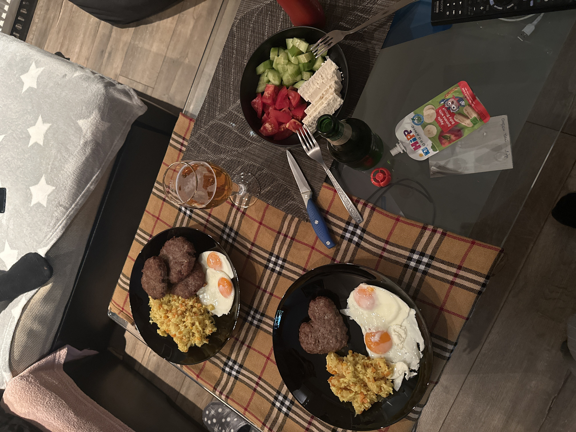 The image captures an indoor scene, possibly a meal setting, on a glass-topped table covered with a checkered brown, red, and black cloth. Two black plates are visible, each containing what appears to be a patty, fried eggs, and a scoop of yellow, rice-like dish. A stemmed glass with beer sits next to one of the plates.
Above these plates, a black bowl holds a salad composed of diced cucumbers, chopped tomatoes, and blocks of what seems to be feta cheese. Cutlery, including forks and a knife with a blue handle, are arranged near the bowl.
On the glass part of the table, a squeezable pouch labeled "Hipp Organic - Apple Banana with Baby Biscuit" is visible, along with a bottle of beer (possibly a dark lager).
In the background, a sofa with a pillow covered in white stars is partially visible, as is part of a remote control.
The setting is likely a home. There is no information to suggest that this photo was taken in the city of Burgas, Bulgaria.