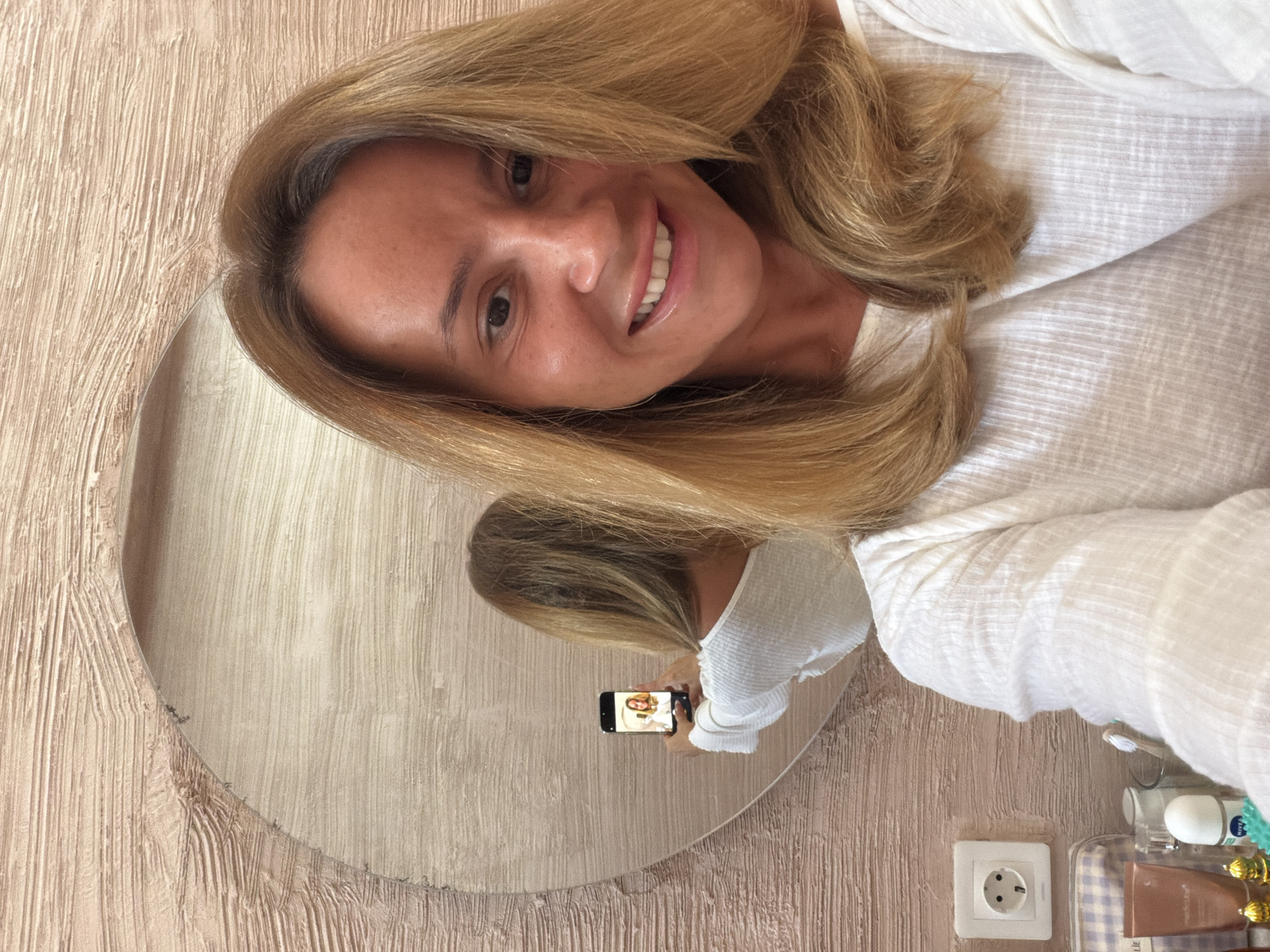 A woman with shoulder-length blonde hair is smiling and looking at the camera, taking a selfie in a circular mirror. The mirror is mounted on a textured, light brown wall. Reflected in the mirror is a view of the woman holding a smartphone, also displaying her image on the screen. To the right of the mirror, on a surface or shelf, are toiletries and personal care products. Below the mirror, a European-style electrical outlet is visible on the wall. The location is possibly a bathroom or dressing area. The context of the image does not provide enough information to confirm the location in North Kuta, Indonesia. The scene is well-lit.