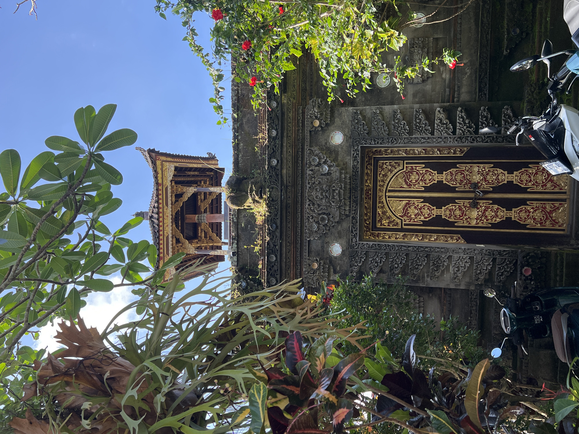 The image is taken in Petulu, Indonesia, showing a building with a traditional Balinese style entrance, visible between lush tropical vegetation. The entrance has an ornately carved gray stone frame around an intricately decorated dark brown wooden door with gold and red accents. A motorcycle is parked partially obscured by foliage to the right of the door. Above, red hibiscus flowers grow from vines cascading down the wall. A small tower structure with golden embellishments can be seen to the left, peeking over the top of the gate. The sky is blue and clear in the background.