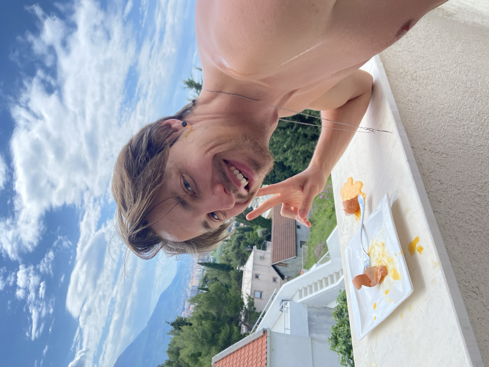 A shirtless man with brown hair and a silver necklace, partially visible from the chest up, smiles and holds up a peace sign while standing on a white balcony overlooking a scenic view. The sky is blue with scattered white clouds. Below, a landscape of green trees and buildings stretches towards distant blue mountains. In the lower center of the image, a broken egg lies on a white plate with a fork on the balcony. Based on the architecture and landscape, the location is likely in Šušanj, Montenegro.