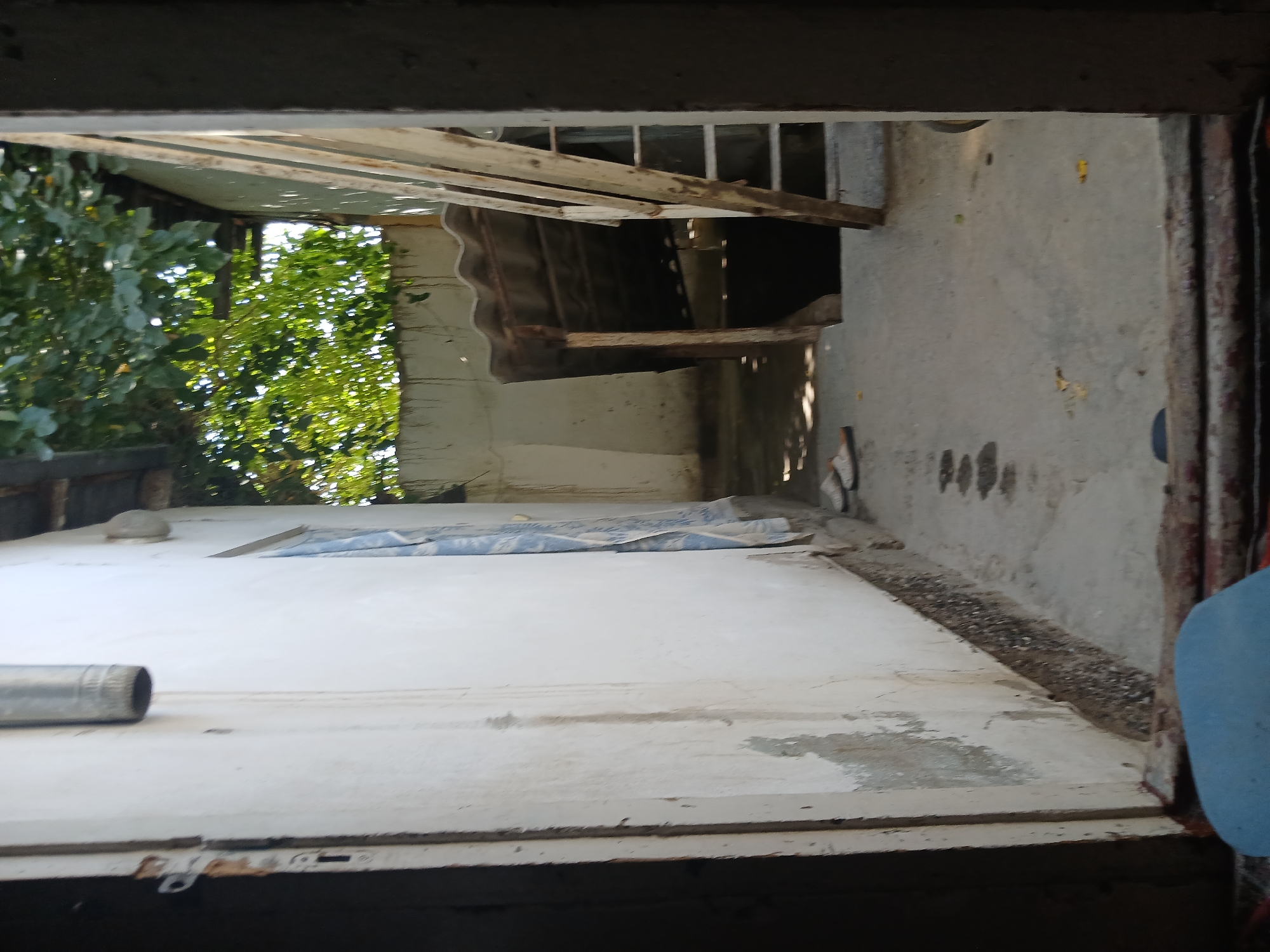 The image shows a narrow outdoor walkway between two buildings. On the left, there is a white wall with a dark doorway. On the right, there is another building with a corrugated roof and a wooden ladder. Green foliage is visible above the roof. The ground is concrete and appears to be stained in several places. Two pairs of sandals are on the ground near the entrance to the building on the right. A metal downspout is attached to the wall on the left. The scene appears to be in a state of disrepair, with peeling paint and debris on the ground.