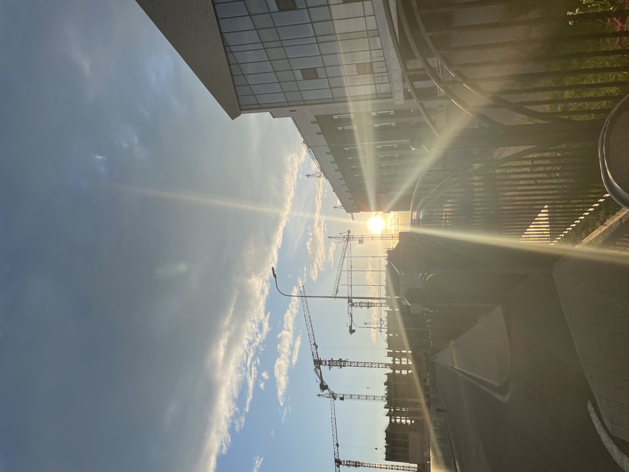 The image depicts a construction site in Stavropol, Russia, during what appears to be either sunrise or sunset, with the sun prominently visible and casting strong rays of light. Multiple tall construction cranes are erected, silhouetted against the sky, with unfinished buildings visible behind them. The foreground features a paved road and sidewalk, bordered by a metal fence. To the right, a modern building with large glass windows is partially visible, reflecting the sunlight. The sky is partly cloudy, with patches of blue and grey clouds. A lamppost stands near the center of the frame, and a few indistinct figures can be seen walking on the sidewalk. The overall scene conveys a sense of ongoing development or urban construction.