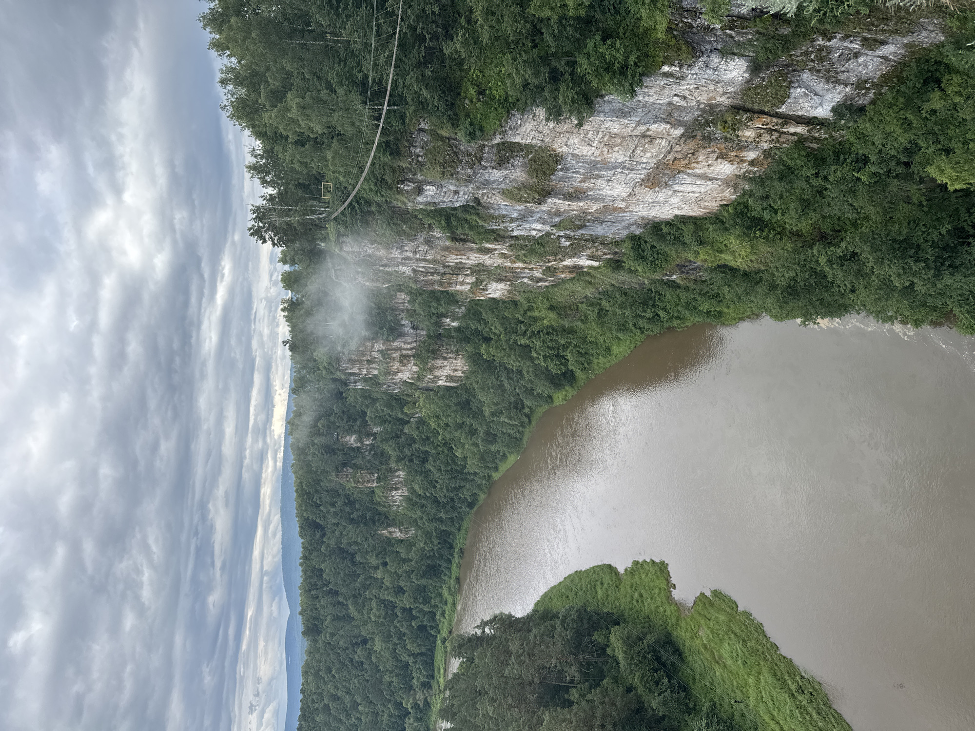 This image shows a sweeping view of a lush green forest bordering a wide, brown river. A sheer, rocky cliff face, partially covered in vegetation, rises from the forest on the left side of the frame. A suspension bridge, made of cables and wooden planks, is visible spanning across the cliff and forest. A plume of mist or fog rises from the cliff face near the bridge, suggesting a waterfall or humid conditions. The sky is overcast with heavy, grey clouds, indicating a cloudy or possibly rainy day. The light is diffused and the overall atmosphere appears serene and natural. The photo is taken from a high vantage point, looking down and across the river valley.
