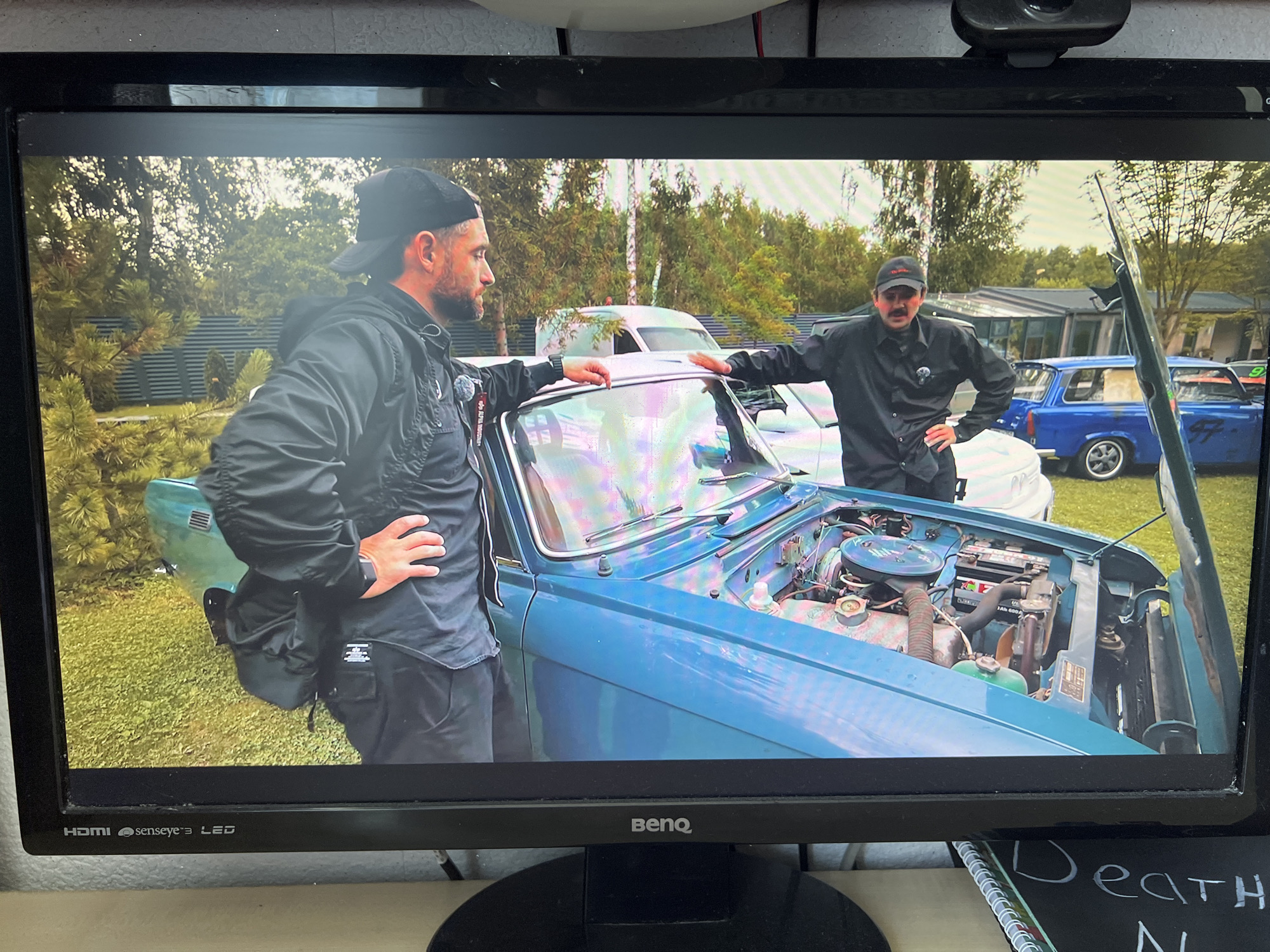 The image shows a computer monitor displaying a video. On the screen, two men stand next to a teal-colored vintage car with its hood open, revealing the engine. The man on the left is wearing a black jacket and a black baseball cap, and he has his left hand on the car's hood. The man on the right is wearing a black collared shirt and a black baseball cap, with a mustache. He has his right hand on his hip and his left hand on the car's hood. The background shows trees, a fence, and other vehicles, including a blue car. A notebook with the words "Death N" is visible in the lower right corner of the image, outside the monitor's display. The monitor itself is a BenQ LED model. The overall scene appears to be outdoors, possibly at a car gathering or event. No specific location in the city of Kanash, Russia is identifiable from the image.