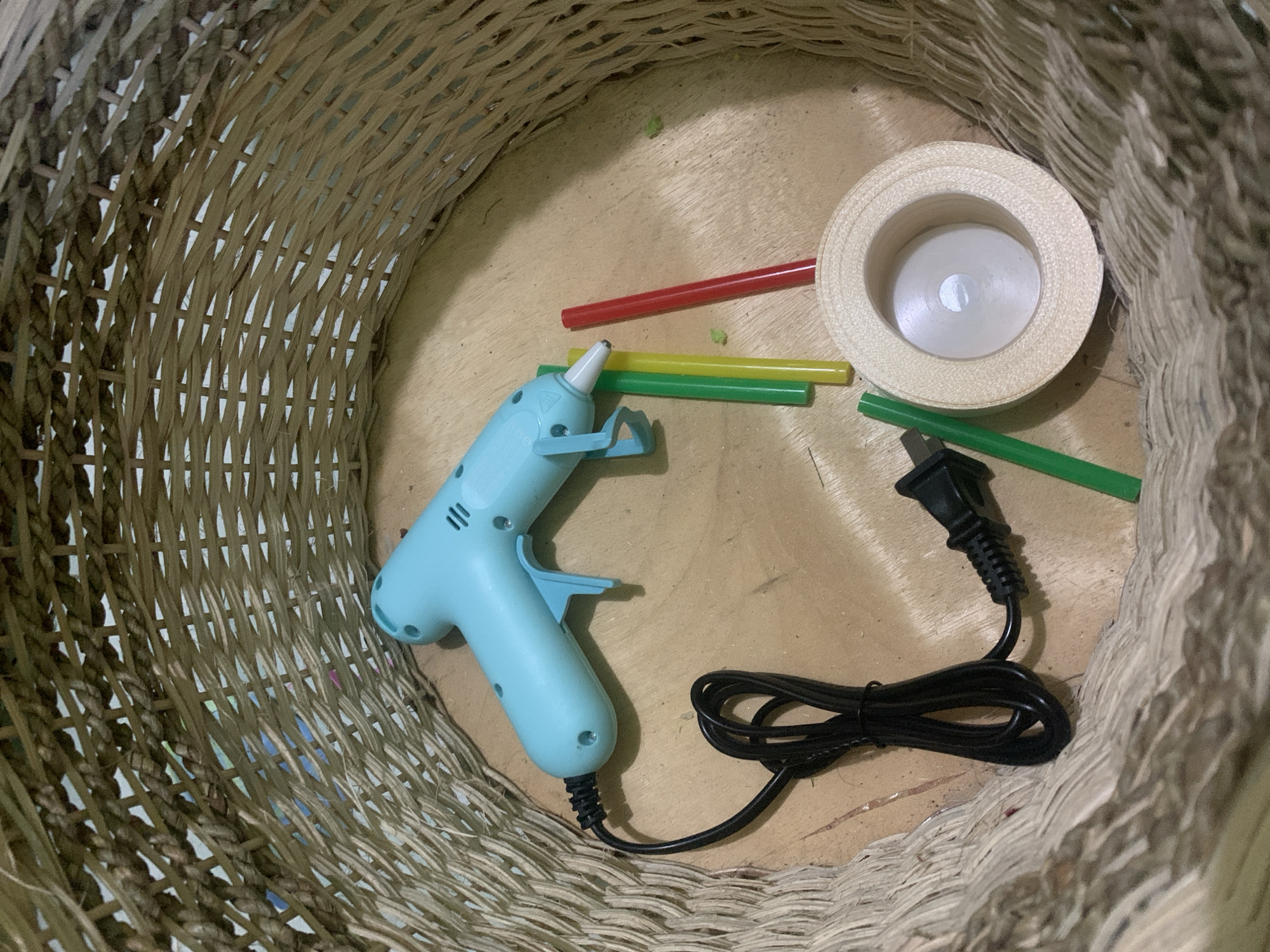A light blue hot glue gun with a black cord and plug rests on a wooden surface inside a woven basket. Three glue sticks, two green and one yellow, are positioned to the right of the glue gun, with a red glue stick slightly above them. A roll of light beige ribbon sits to the right of the green and yellow glue sticks. The woven basket is made of natural, light-brown fibers and has a wide opening. The image appears to be taken in indoor lighting. There is no indication of the specific location in Kuala Pembuang, Indonesia, or any people present.