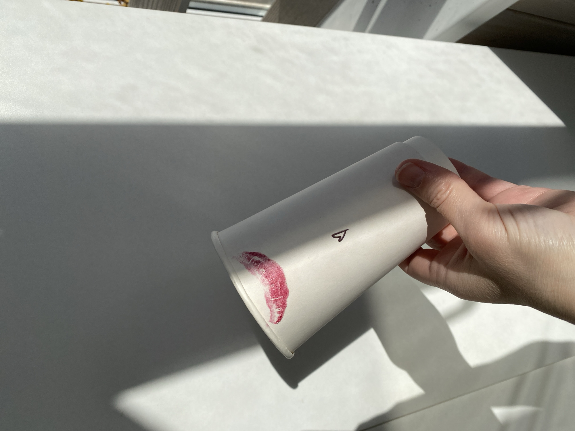 A hand holds a white paper cup with a lipstick kiss on the rim and a drawn heart. The scene is lit by sunlight, creating shadows on a white surface. The location is unknown, although the user specified Chaikovsky, Russia.