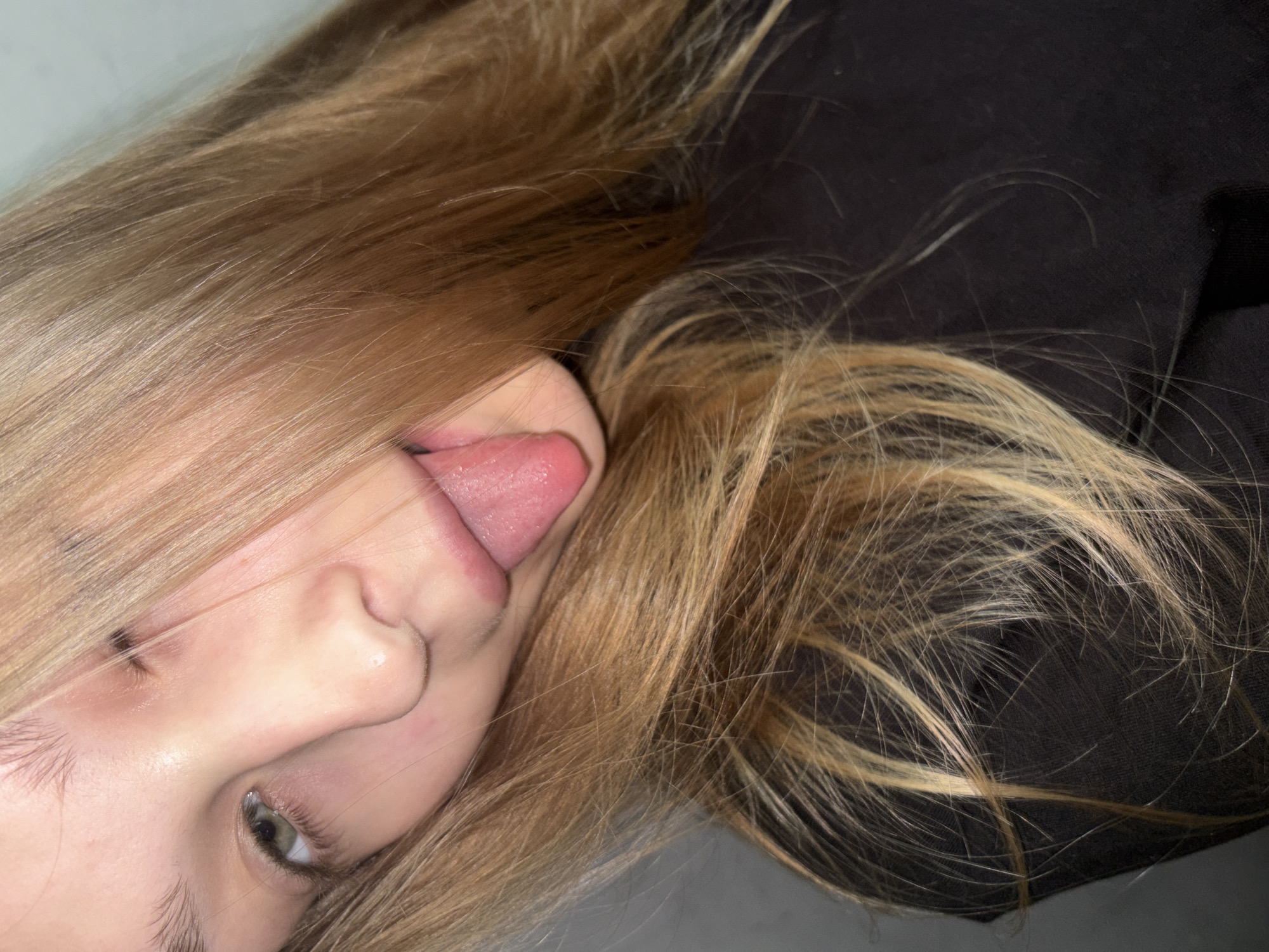 A light-skinned individual, appearing to be a young woman, is depicted in a close-up, angled portrait, looking directly towards the camera. Her head is tilted, and her tongue is extended outward from her mouth, appearing vibrant pink and moist. Her long, straight hair, which is blonde or light brown with lighter highlights, falls across the right side of her face, partially obscuring her right eye. Her left eye is visible, showing a light green or hazel iris. Her complexion is fair. She is wearing a plain black garment, likely a shirt or top, visible across her shoulder and chest area. The background is an indistinct, light-colored, out-of-focus surface, suggesting an indoor setting. No discernible features within the image indicate a specific geographical location, such as the city of Chaikovsky, Russia, or any other region.