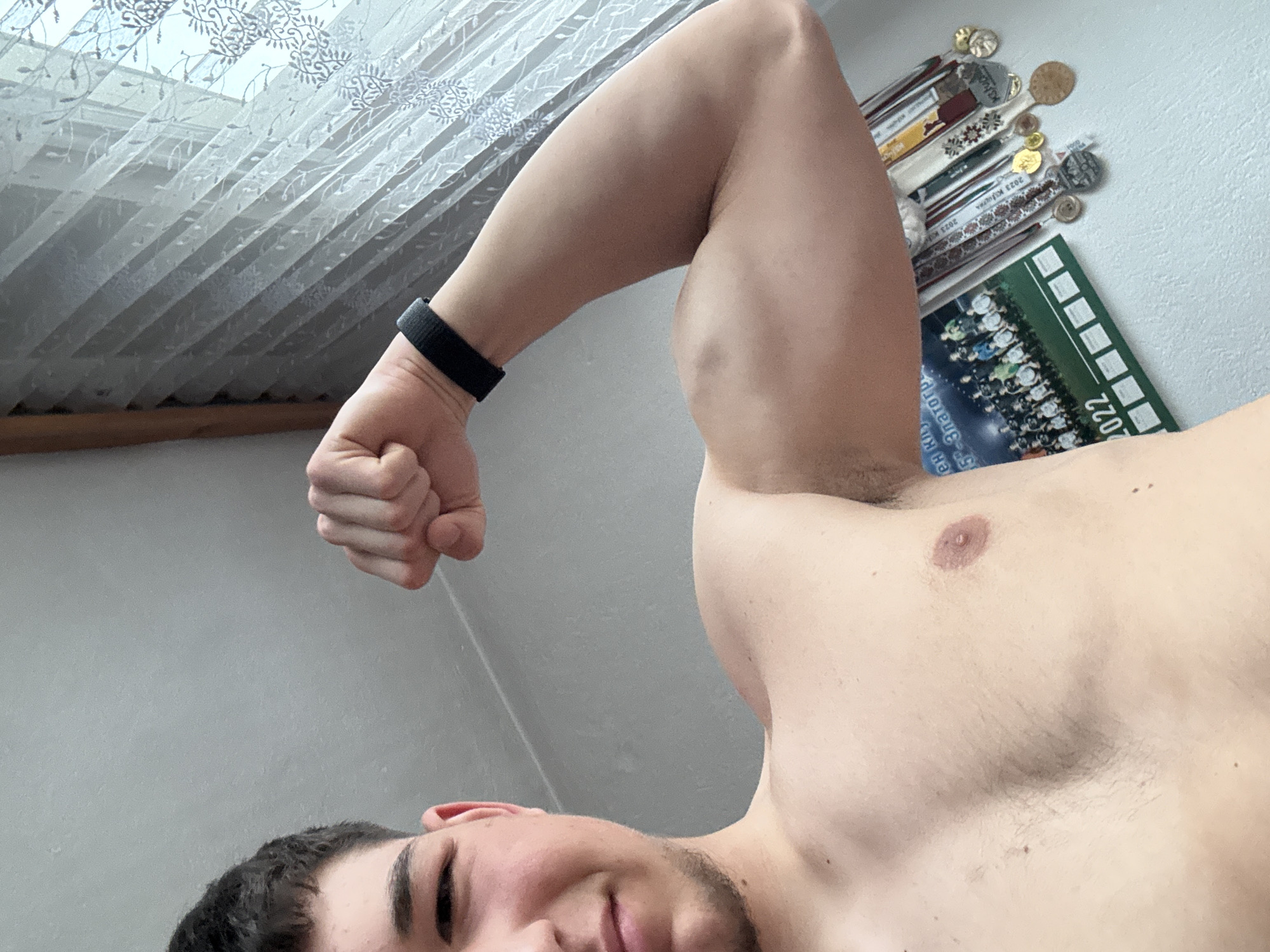 A young man is depicted in a close-up, low-angle selfie, flexing his right bicep. He is shirtless, displaying a muscular physique, with his head partially visible in the bottom left, looking towards the camera with a slight, confident smile. His dark hair and light stubble are visible. A black wristband is worn on his right wrist, and his right fist is clenched as he showcases his arm.
The setting is an indoor room, likely in Zlatograd, Bulgaria. In the background to the left, a large window is covered with white, translucent curtains featuring a repeating floral or leafy pattern, allowing bright, diffused natural light into the space. On the right, against a light-colored wall, there is a collection of awards or decorative items, some resembling medals or commemorative spoons with intricate designs and ribbons. Below these, a calendar is propped up, clearly displaying "2022" and showing a group photo of people. Visible Cyrillic text on the calendar includes "2022 КАЛЕНДАР" and "ЗЛАТОГРАД". The overall impression is of a casual, well-lit environment.
