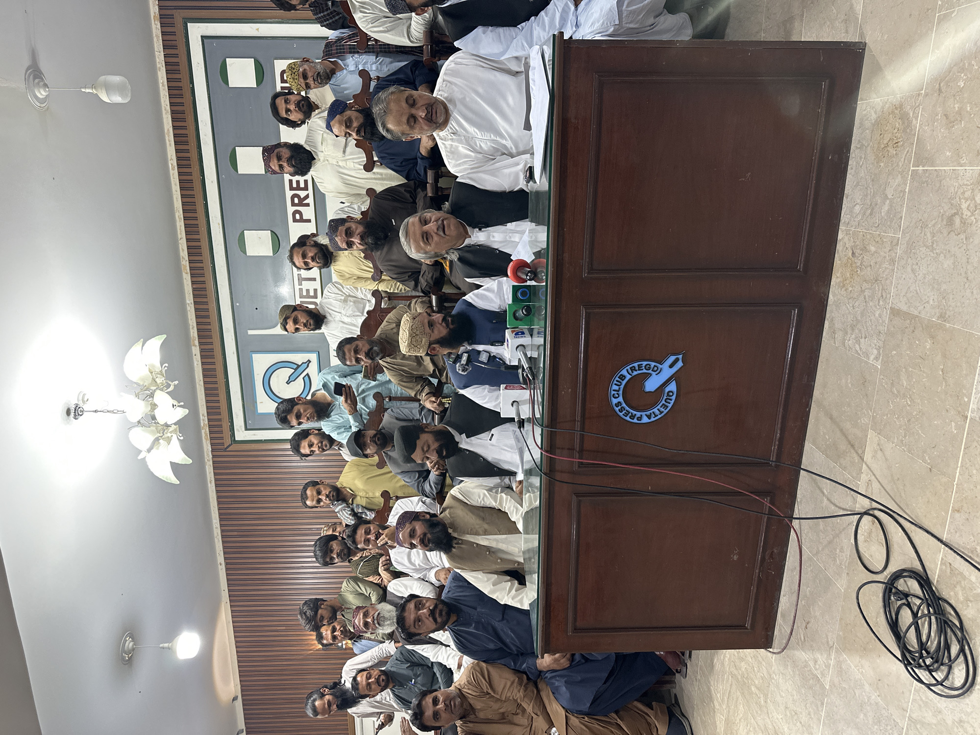 A large group of men are gathered indoors in what appears to be a press conference at the Quetta Press Club (REGD) in Quetta City Tehsil, Pakistan. The men are mostly sitting behind a wooden desk with microphones on top. At least two men are standing and speaking, with one wearing a white shirt. Many men in the audience are wearing traditional Pakistani clothing, including various head coverings. A sign with the words "QUETTA PRESS CLUB (REGD)" is visible on the front of the desk. Cables and wires are visible coming from the desk and microphones. The room has light-colored striped walls and a patterned tile floor. A chandelier hangs from the ceiling in the upper left of the image.