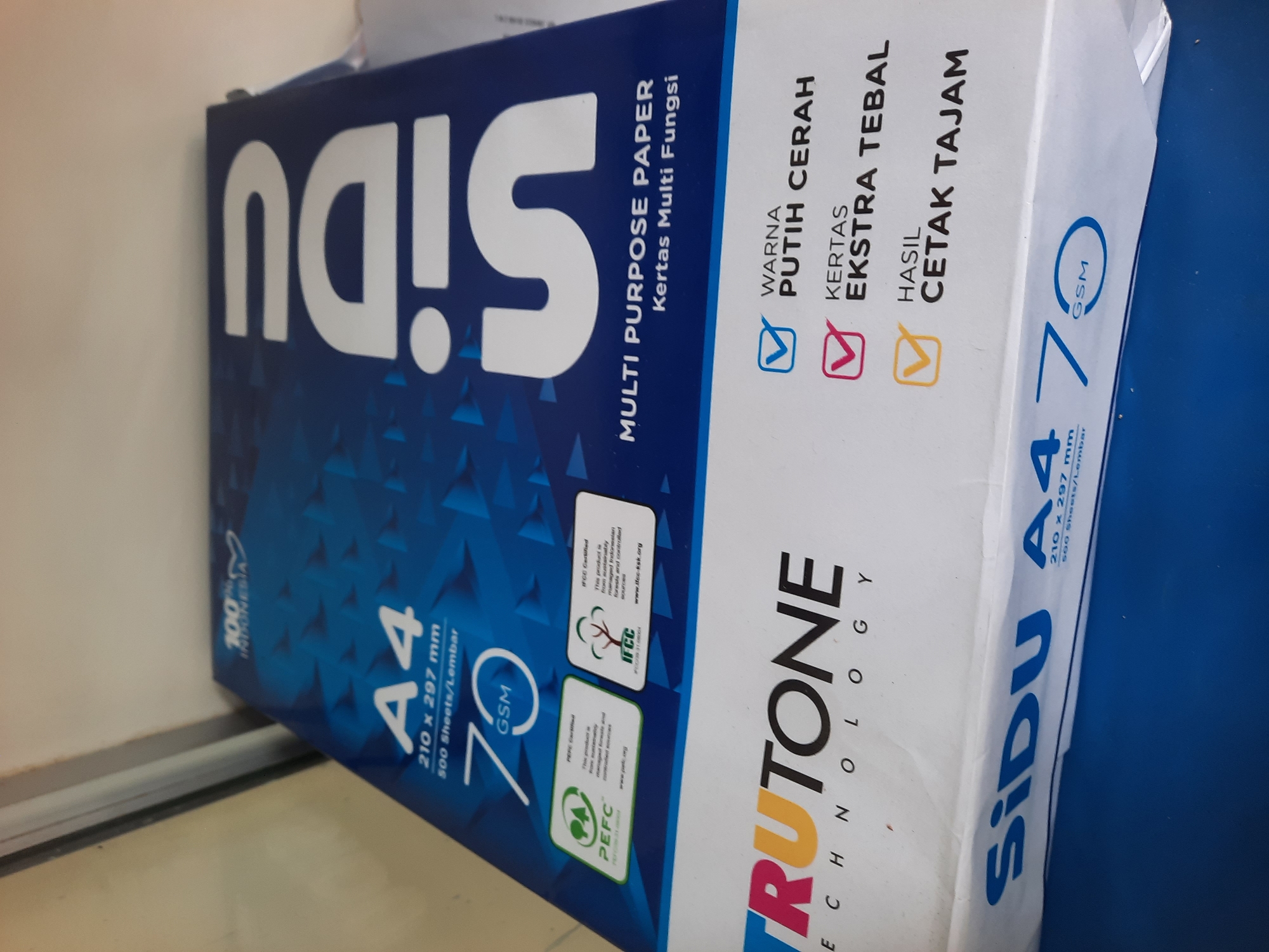 The image displays packaging for Sidu A4 multi-purpose paper. The packaging is predominantly blue with white lettering. Prominently featured are the brand name "Sidu" and the paper size "A4" along with specifications like "210 x 297 mm" and "70 GSM". There are also logos indicating PEFC and IFCC certification. Below the Sidu branding, there is another brand name "TRUTONE TECHNOLOGY" in colorful letters. Text on the packaging highlights features like "WARNA PUTIH CERAH" (Bright White Color), "KERTAS EKSTRA TEBAL" (Extra Thick Paper), and "HASIL CETAK TAJAM" (Sharp Print Result). The items are placed on a blue surface, likely a table or desk, against a neutral background that includes a portion of a wall and a window frame. There are no people visible in the image. The overall context suggests a retail or office setting where paper products are stored or displayed. The location is specified as Lhokseumawe, Indonesia.