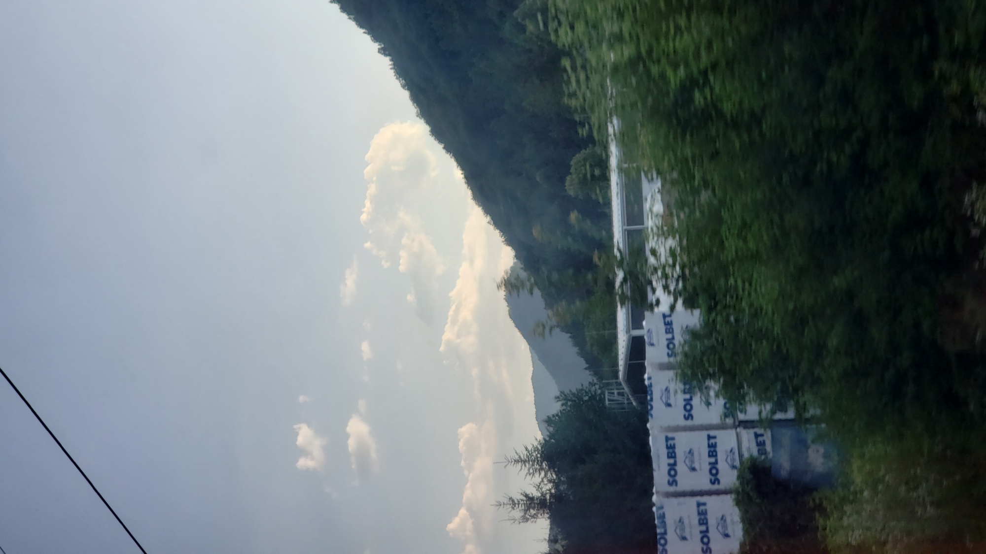 The image depicts a landscape scene in the vicinity of Ľubochňa, Slovakia. In the foreground, stacks of white building materials, possibly insulation boards, are visible. These materials are marked with the brand name "SOLBET". Adjacent to these stacks are dense green trees and foliage. In the midground, there is a white building structure with a roof, partially obscured by trees. The background features rolling hills and mountains covered in dark green forests, under a pale blue sky with scattered white clouds. A thin black line, likely a power line, stretches across the upper left portion of the image. The overall impression is one of a rural or industrial area bordering a natural mountainous environment.