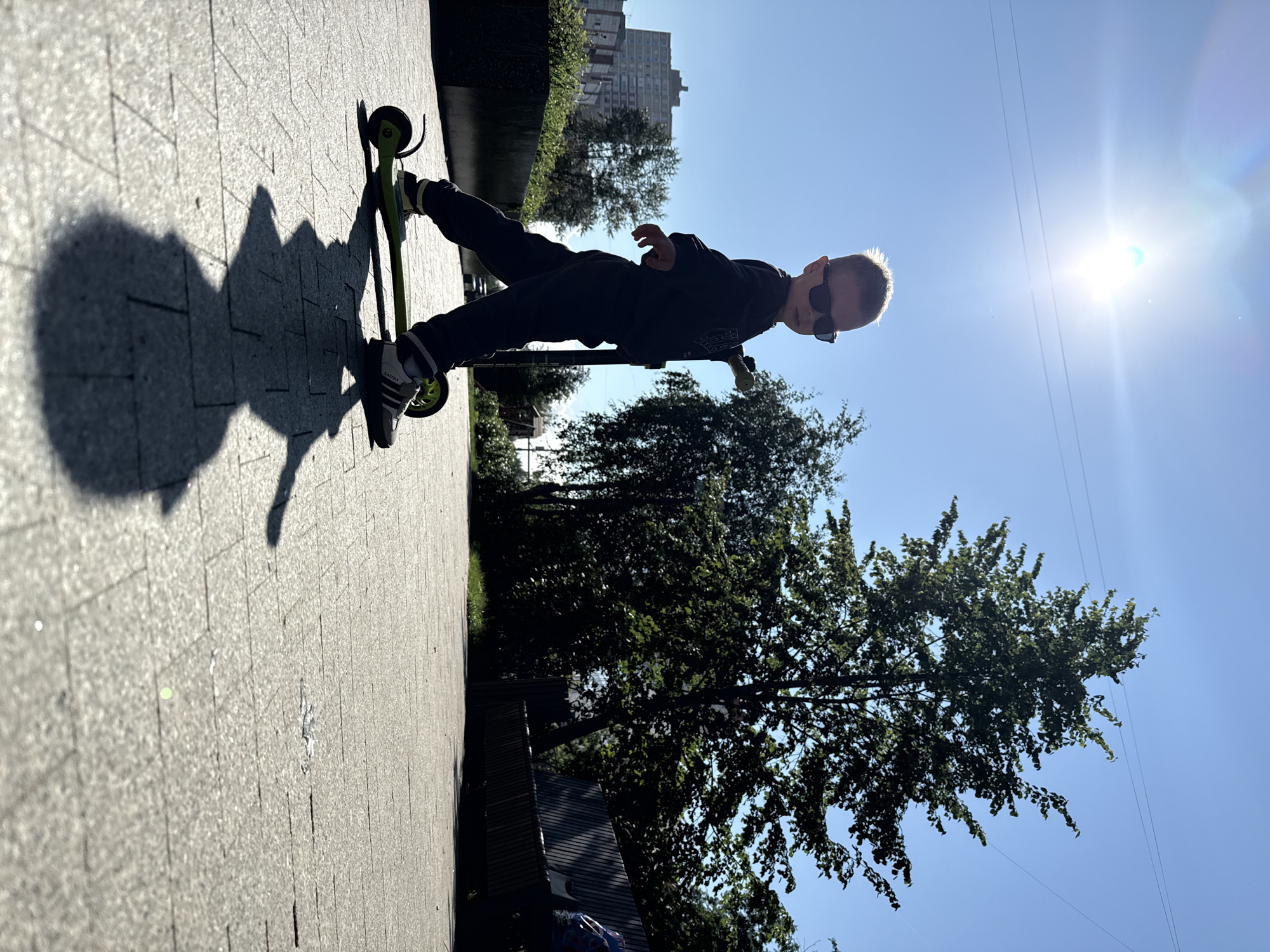A young boy wearing sunglasses rides a green scooter on a paved walkway under a bright sun and clear blue sky. He is dressed in a dark sweatshirt and pants, with athletic shoes. The boy's shadow is cast long on the ground in front of him. To the right of the walkway is a dense area of trees and a wooden bench. In the background to the left, a tall apartment building is visible, suggesting an urban environment. The scene appears to be outdoors, likely in a park or public space within the city of Perm, Russia, given the architectural style of the building. The sun is high in the sky, indicating it is daytime.