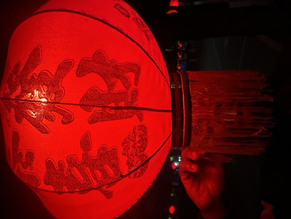 A large, illuminated red Chinese lantern dominates the foreground, centered against a dark, out-of-focus background. The spherical lantern is adorned with dark red Chinese characters, "恭喜发财," and a stylized decorative element below them. A decorative fringe of multiple red strands hangs from the lantern's base. In the lower-left, a human hand, bathed in the lantern's red light, is observed touching the fringe. The blurred background contains multiple distant red lights, suggesting additional lanterns or light sources along a street or public area at night in the city of Jiangjin, China.