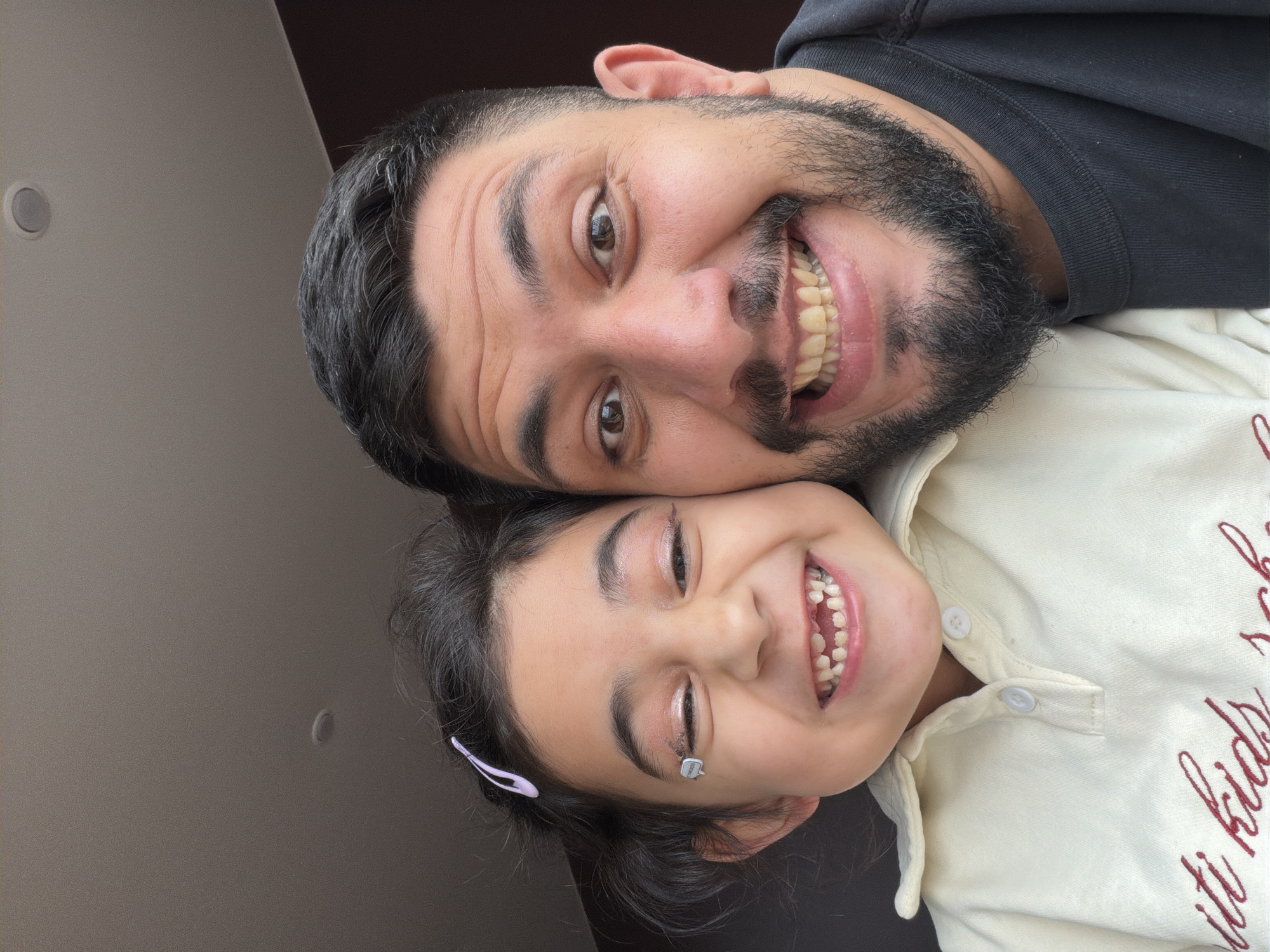 A man and a young girl are posing for a close-up photo. The man is older, with dark hair and a full beard, wearing a black hooded shirt. The girl is younger, with dark hair and a light-colored shirt with red writing. Both are smiling broadly, showing their teeth. The background is a plain wall with a single light fixture visible. The photograph is taken from a slightly angled perspective, with the subjects tilted relative to the frame. There is no visible information to indicate that the photo was taken in Krasnoyarsk, Russia.