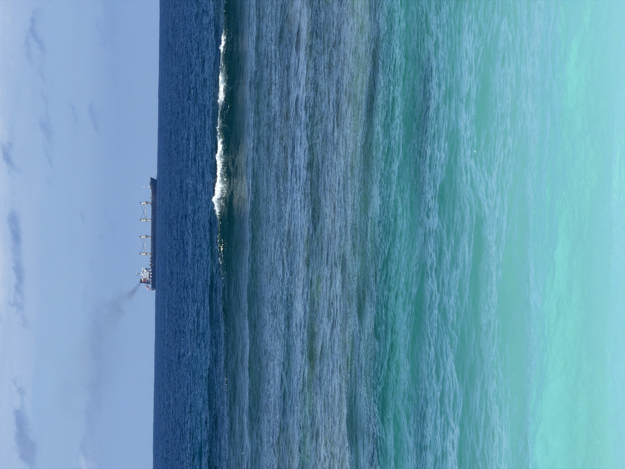 A vertical shot shows a view of the ocean near Ungasan, Indonesia. The left side features a blue sky with scattered clouds and a dark blue ocean with a cargo ship emitting smoke. A foamy white wave line marks the edge of the darker ocean. The water gradually transitions to lighter shades of blue and turquoise towards the right side of the image, showing the ocean's color gradient.