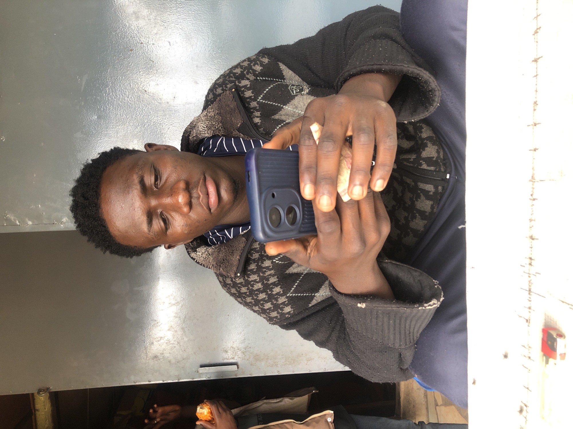 A young Black man is sitting and looking at his cell phone. He is wearing a black sweater with a grey patterned design and a blue and white striped shirt underneath. He is also wearing navy blue pants. He is holding a dark blue cell phone in his hands. In the background, there is a grey metal door. To the left of the man, another person is partially visible, holding a piece of food. A white surface with some markings and a small red object is in the foreground. The image appears to be taken outdoors, possibly in an urban setting.