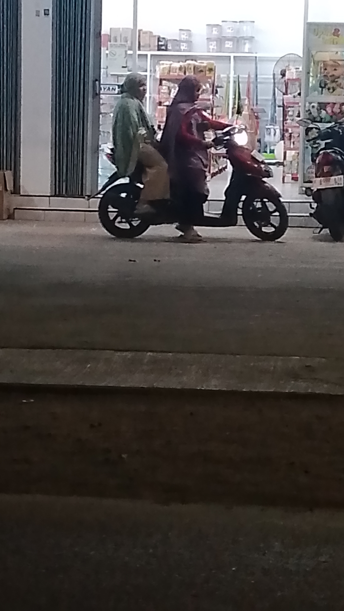 Two individuals are riding on a red motorcycle in front of a building with a corrugated metal facade. The person in the front, who appears to be driving, is wearing a dark headscarf and a maroon jacket. The passenger behind them is wearing a light green hijab and a light brown outfit. They are both facing forward, suggesting they are moving. The background shows shelves filled with products, indicating a retail environment. The motorcycle is on an asphalt surface, and the scene appears to be outdoors, possibly on a street in Karanglangu, Indonesia, given the context.