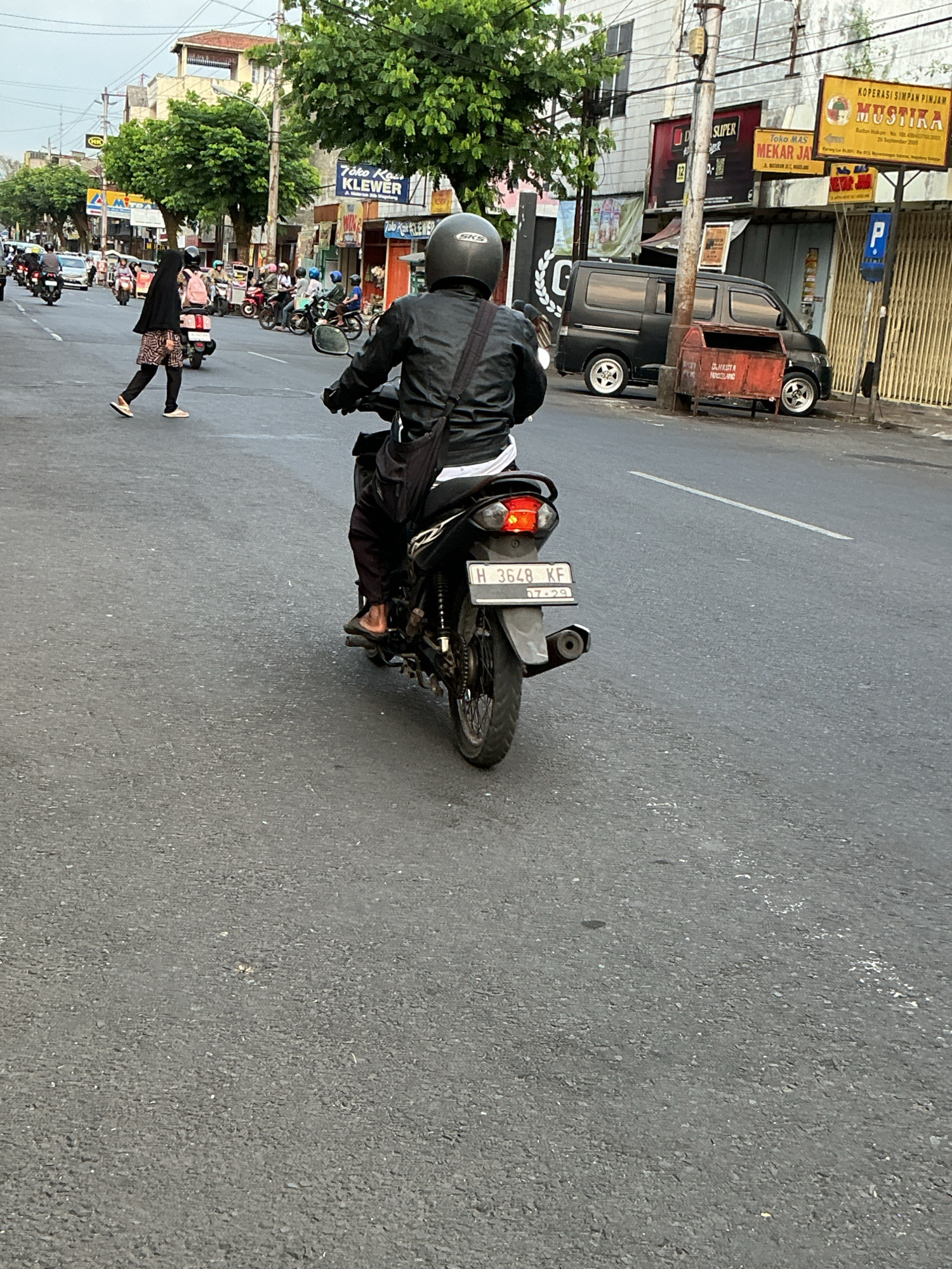 The image captures a street scene in Magelang, Indonesia. A person is riding a motorcycle in the foreground, viewed from behind. The rider wears a black leather jacket and a black helmet. The motorcycle has a license plate that reads "H 3648 KF" with "07-29" below it. In the background, several other motorcycles and a black van are visible. A woman in a black headscarf and patterned skirt is crossing the street. Buildings with signs, including "Joko Mas Mekar Jaya" and "KOPERASI SIMPAN PINJAM MUSTIKA," line the street. Trees and utility poles are also present. The road is paved and appears to be an urban thoroughfare.