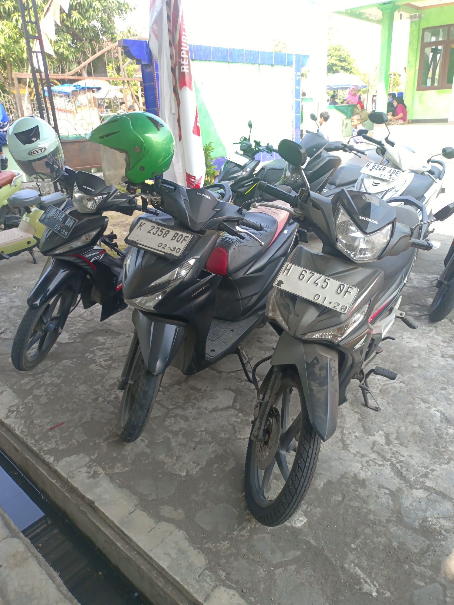 The image captures a street scene in Sarimulyo, Indonesia, dominated by several parked motorcycles. In the foreground, a dark gray Honda motorcycle with the license plate "H 6745 OF" is prominently displayed, its headlight illuminated. To its left, another dark gray Honda motorcycle with the license plate "K 2358 BOF" is also visible. A light green helmet rests on the handlebars of the motorcycle on the left, and a vibrant green helmet is placed on the handlebars of the motorcycle to its right. Further to the left, a portion of a light yellow motorcycle can be seen. The motorcycles are parked on a paved surface, likely a street or sidewalk, with a concrete gutter running along the bottom of the frame. In the background, a building with green and blue accents is present, and a flagpole stands tall. Several individuals, including a woman in a pink hijab and another in a pink shirt, are visible in the middle ground, suggesting a public or communal space. The overall atmosphere appears to be that of a typical day in a bustling Indonesian town.