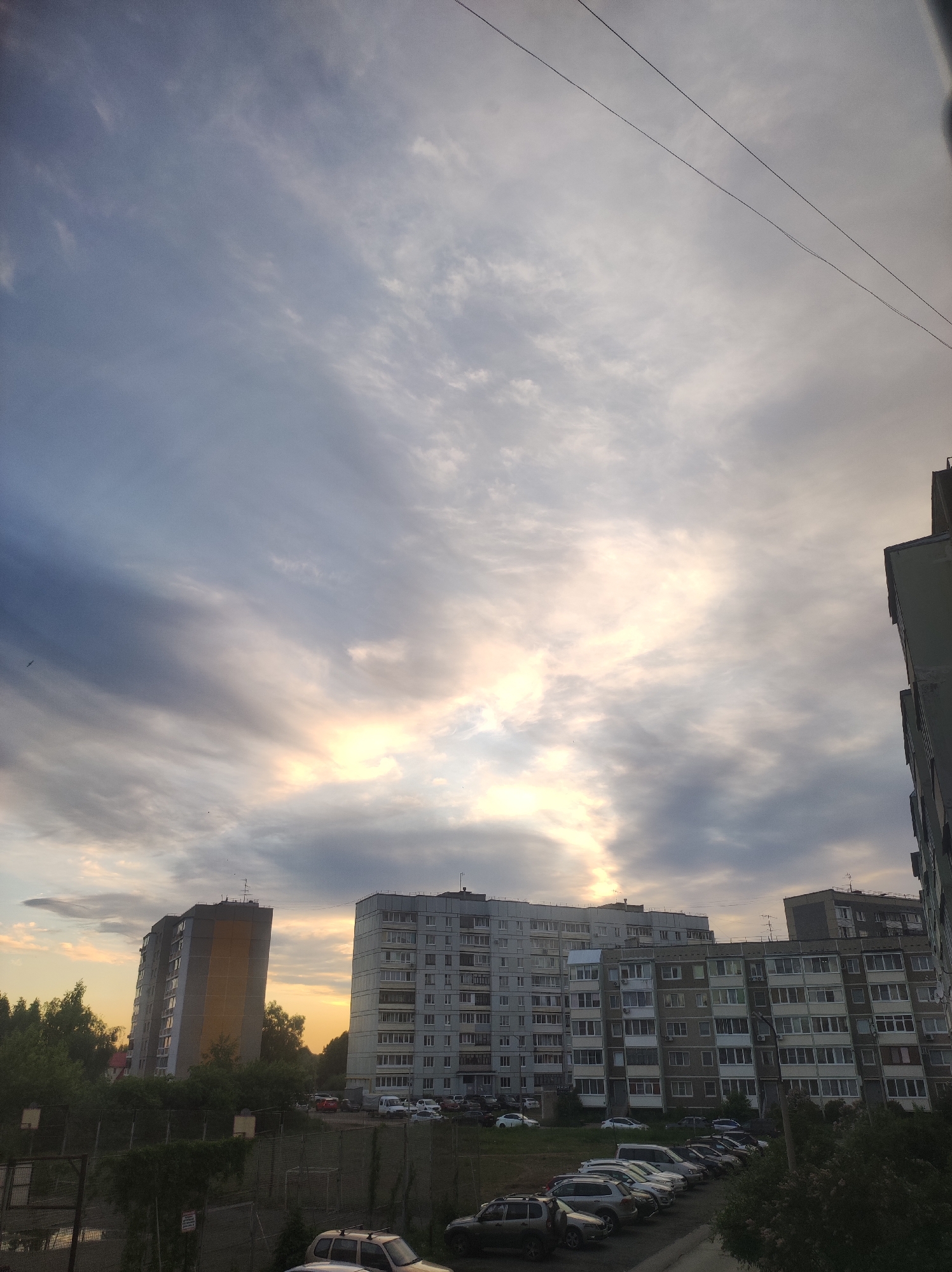The image shows a cloudy sky above residential buildings in Chaikovsky, Russia. Several multi-story apartment buildings are visible, along with parked cars in a lot below. Wires stretch across the top of the frame. Trees and greenery are interspersed throughout the scene. The sky is primarily gray and white with a hint of sunset colors.