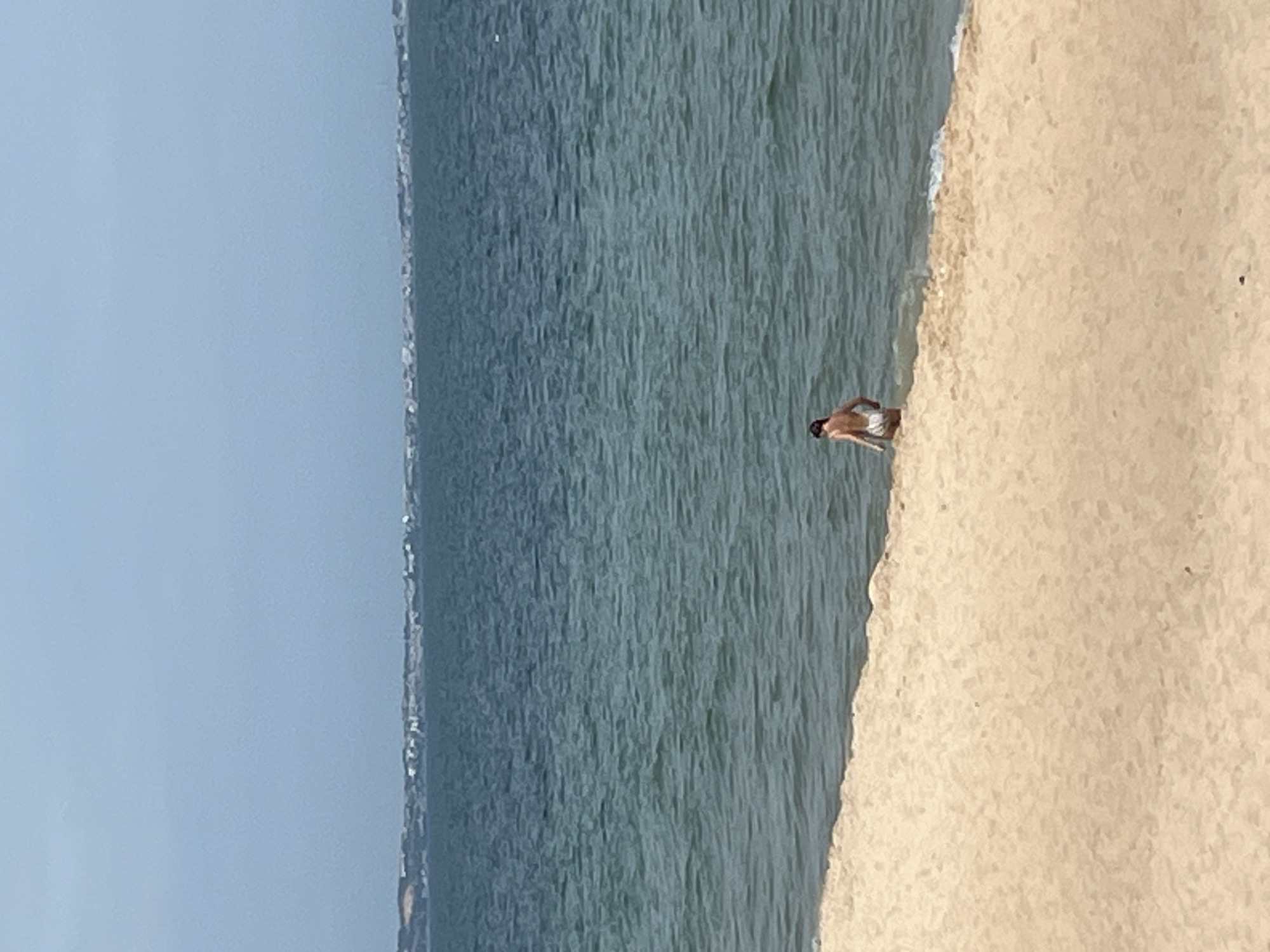 A person wearing white shorts is standing in shallow water at the edge of a sandy beach. The water is a deep blue-green with small waves lapping at the shore. In the background, a distant shoreline is visible with buildings and vegetation. The sky is a pale blue with some haze. The scene depicts a natural environment with a solitary individual engaged in a water activity. The location is not identifiable as Mui Ne, Vietnam.
