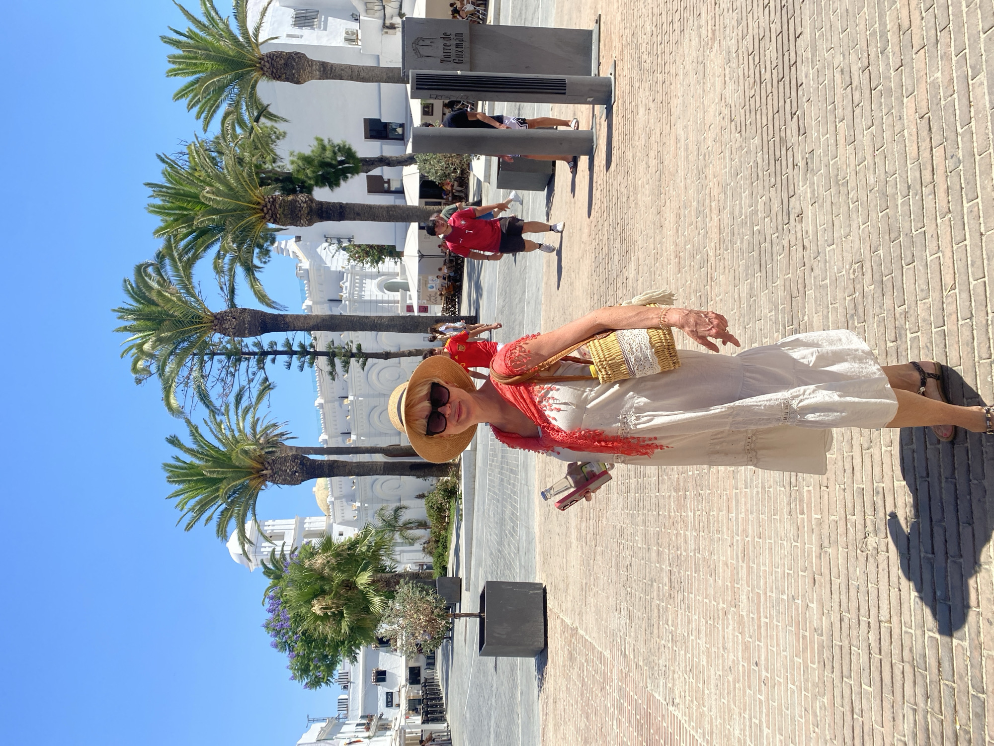 A light-skinned woman with blonde hair and sunglasses, wearing a white tiered dress, a straw hat, and a red scarf, stands in a brick-paved plaza under a bright blue sky. She is holding a bottle of water and a phone. In the background, palm trees and white buildings with arched windows and a dome are visible. Several people are walking in the plaza, and outdoor seating areas with umbrellas are set up in front of some buildings. A sign reads "Torre de Guzman." The scene appears to be daytime in a warm climate, likely the town of Conil De La Frontera, Spain, given the architecture and signage.