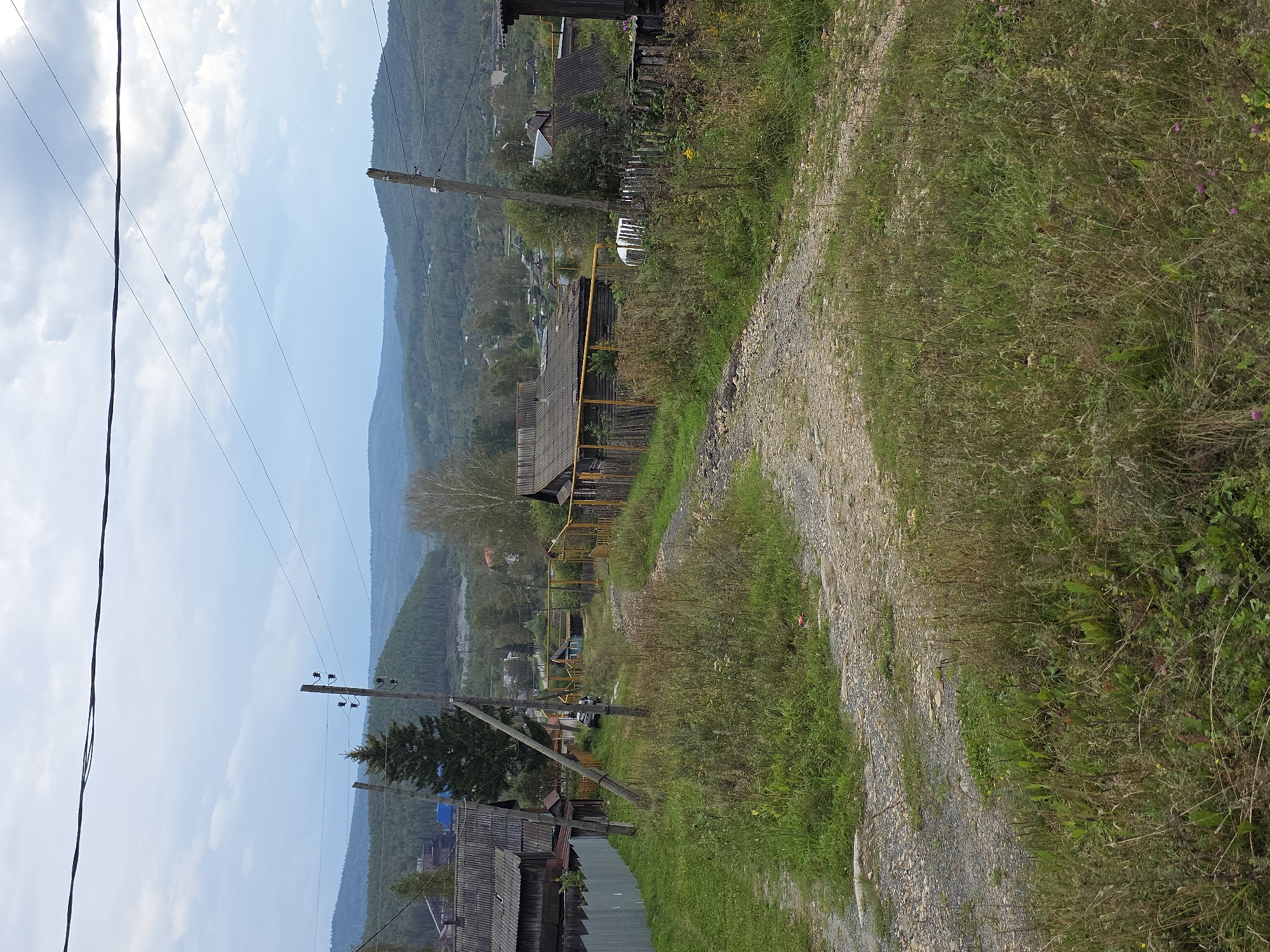 A dirt road winds through a grassy, somewhat overgrown area in a rural settlement. The road is surrounded by wild grasses and some wildflowers. Several wooden houses with corrugated roofs are visible, some with wooden fences. A yellow pipeline runs along the fence of one of the houses. Utility poles with wires are present, indicating electricity infrastructure. In the background, rolling, forested hills rise under a cloudy sky. The scene appears to be a residential area within or on the outskirts of Magnitka, Russia. There are no people visible in the image.